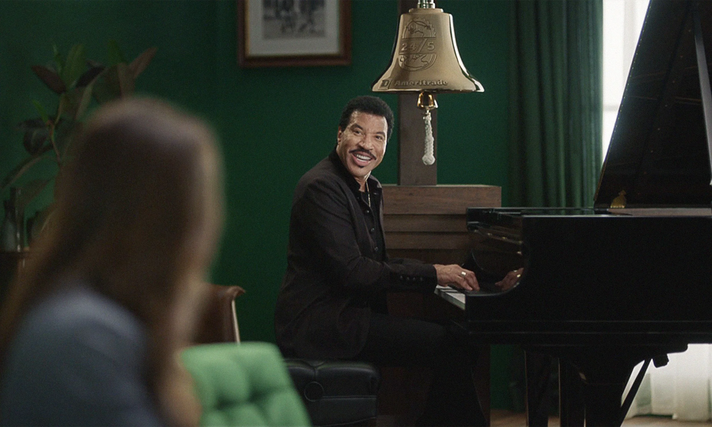 A man playing a piano and smiling, with a woman in the foreground, in a room with green walls and a window with curtains.