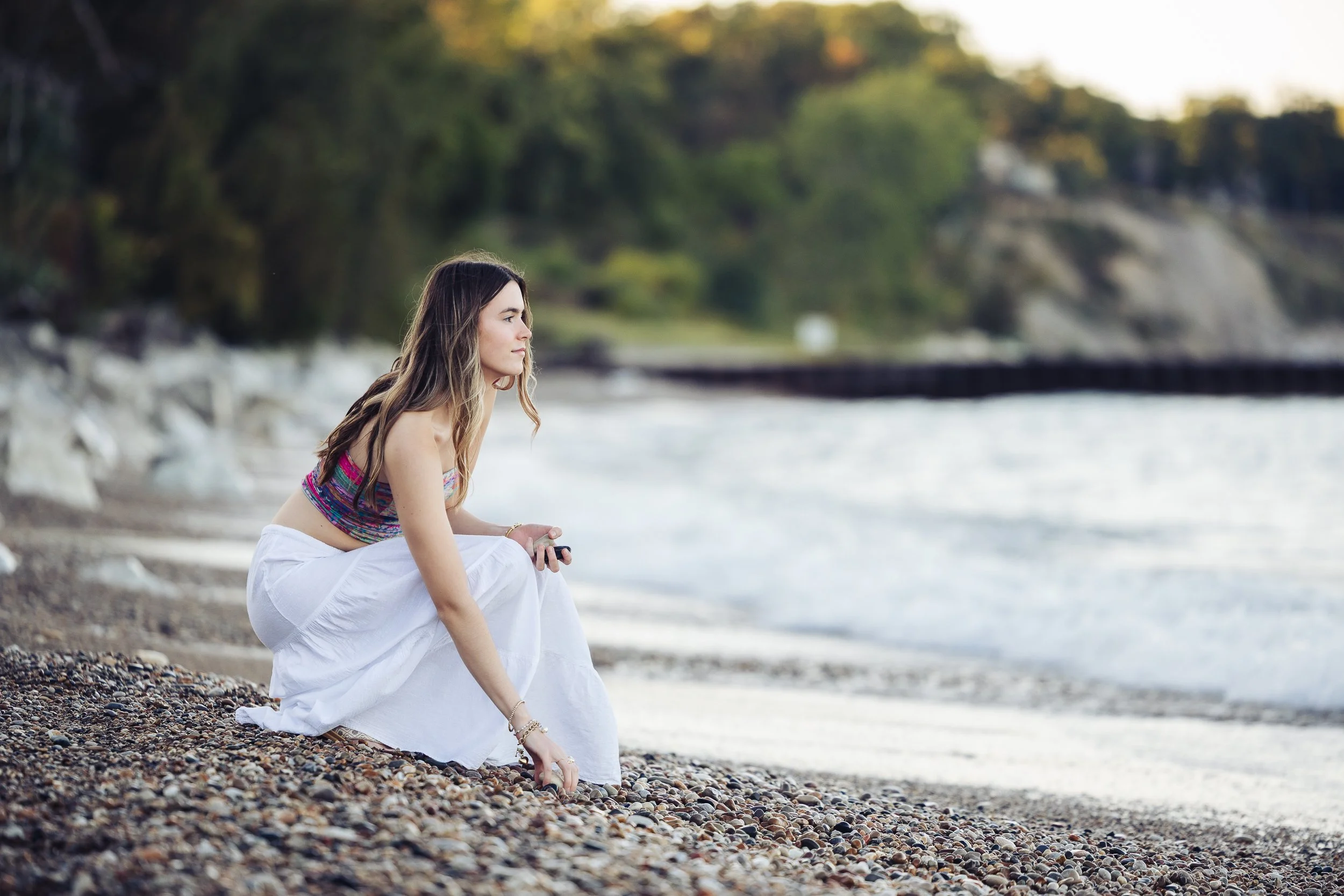 teen girl crouching on a stone beach looking at the water, holding rocks in her hand