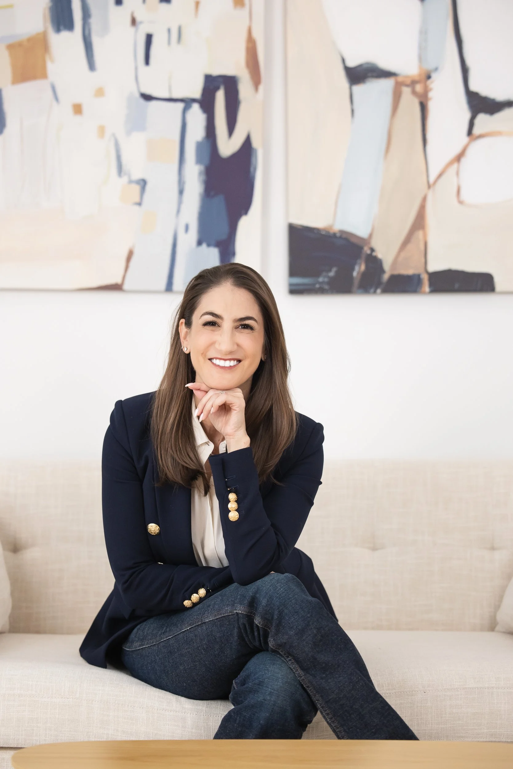 woman seated on a white couch, legs crossed, smiling at the camera