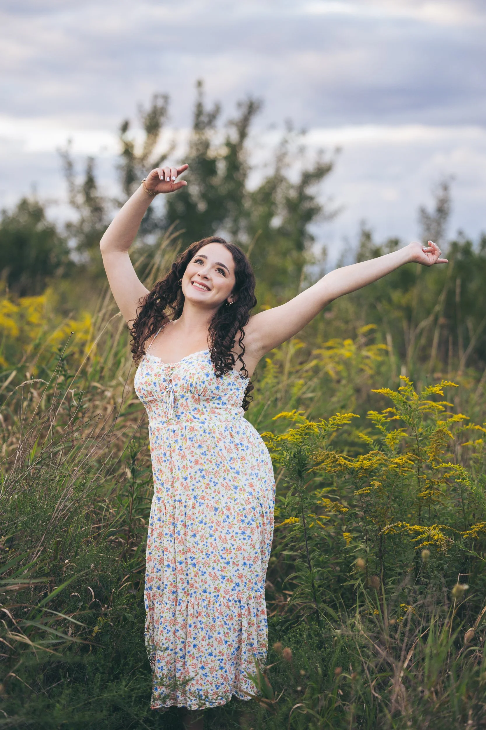 teen girl in a prairie field dancing with hands waving above her head, a smile on her face, wearing a flower print sundress