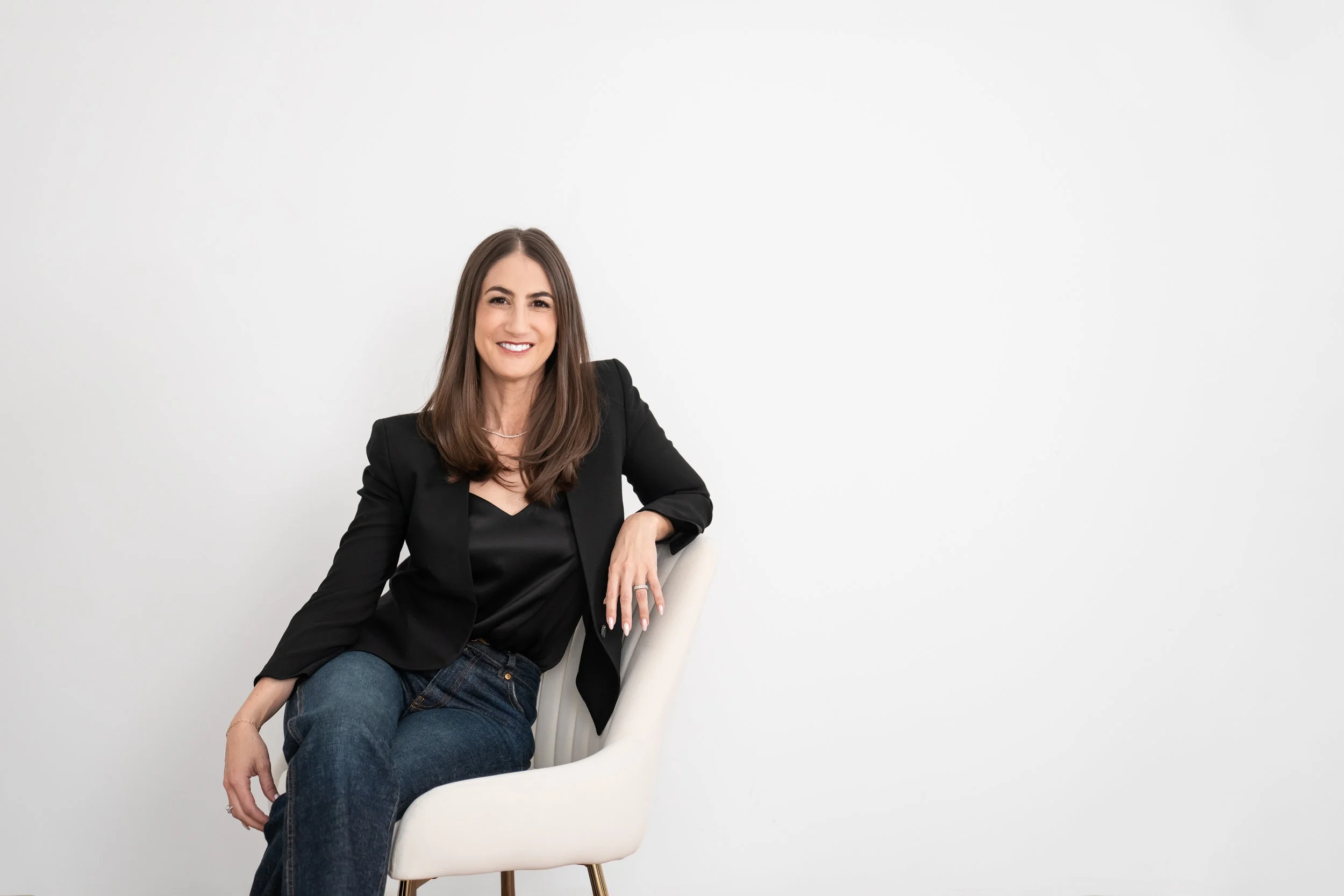 woman seated on a white chair, with a white background, smiling at the camera