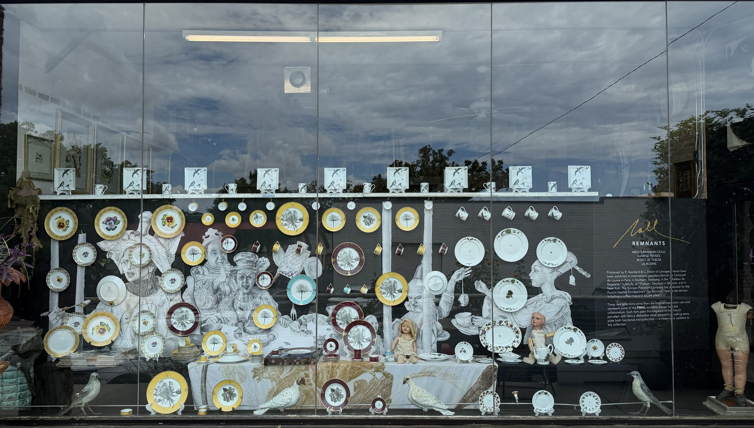 Storefront window display with vintage and collectible items, including plates, mugs, and figurines, featuring floral and animal designs, and a black informational sign about the collection.