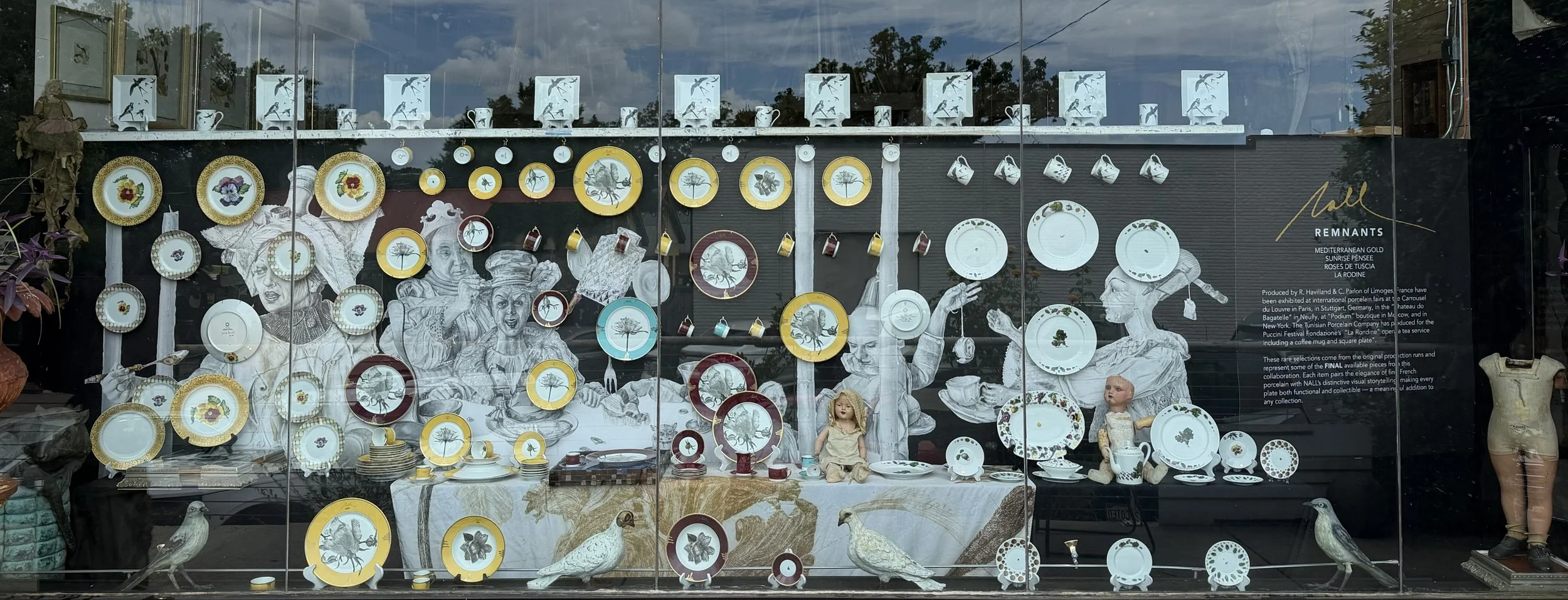 Display window showcasing decorative porcelain plates and cups with botanical and bird motifs, arranged with dolls, bird sculptures, and informational text about the collection.