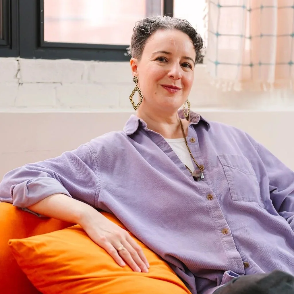 A photo of a white, non-binary person with short, dark, curly hair wearing a purple shirt and funky earrings sitting on an orange sofa