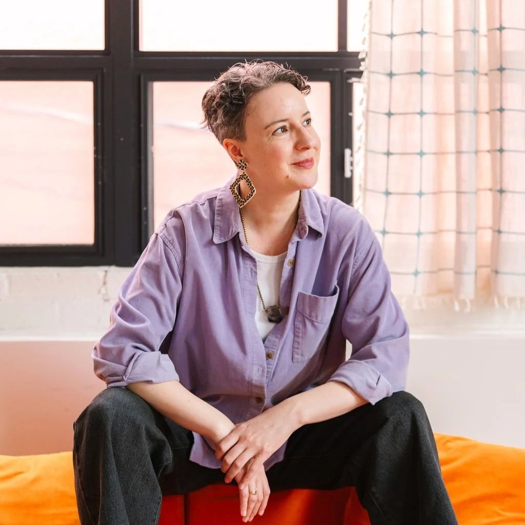Photo of Or, a white non-binary person with short curly dark hair wearing a purple collared shirt and funky earrings, looking off the the side with a slight smile