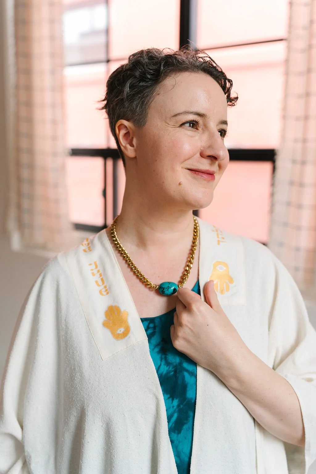 Photo of a white non-binary person with short curly hair looking off to the distance, wearing a turquoise top and white and gold Tallit.
