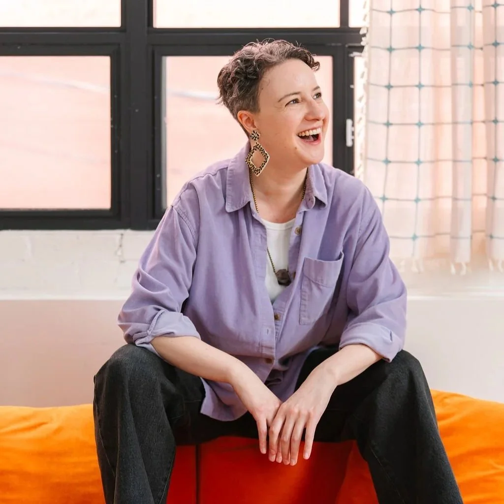 Image of a white, non-binary person with short curly dark hair wearing a purple collared shirt and funky earrings, looking off into the distance and laughing