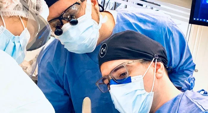 Tres profesionales médicos en un quirófano o área médica, usando batas, mascarillas y gafas protectoras, concentrados en un procedimiento.