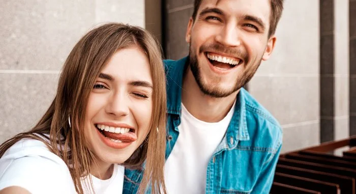 Dos personas sonriendo y posando para la foto, una mujer y un hombre en primer plano, con fondo de pared de ladrillos