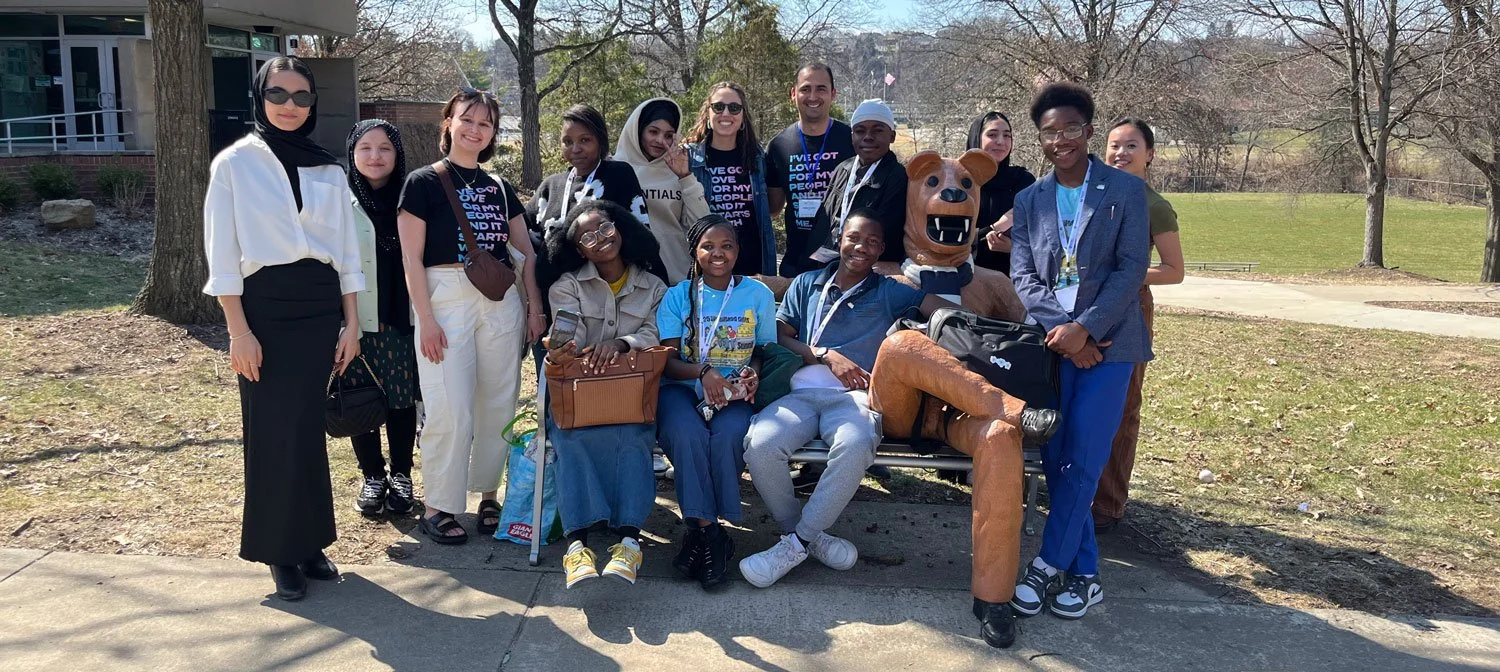 A group of diverse young people posing outdoors on a sunny day. Some are sitting on a bench, others standing. The background features trees and a grassy area.