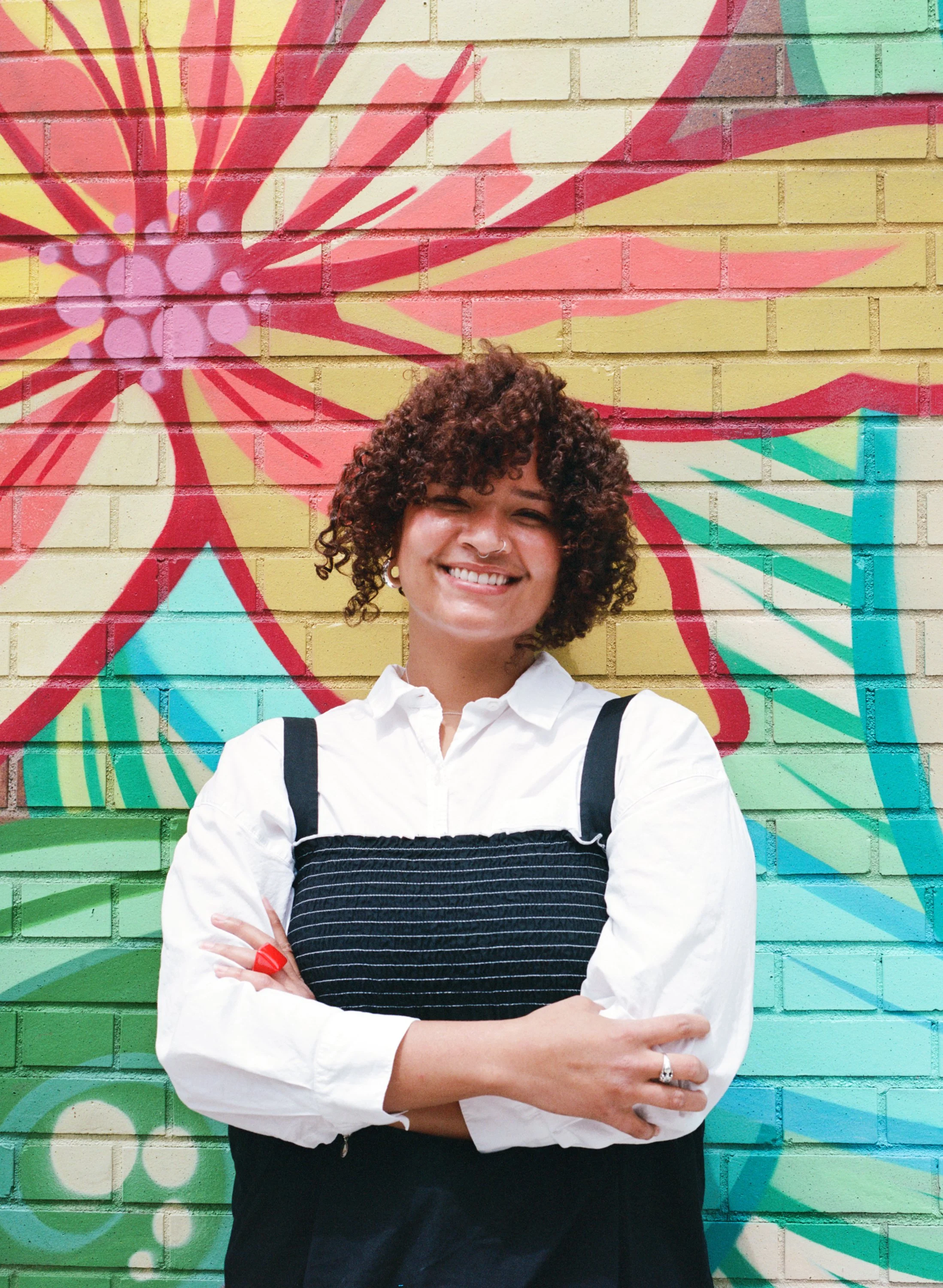 A young woman with curly hair smiling and crossing her arms, wearing a brown plaid shirt against a plain white background.