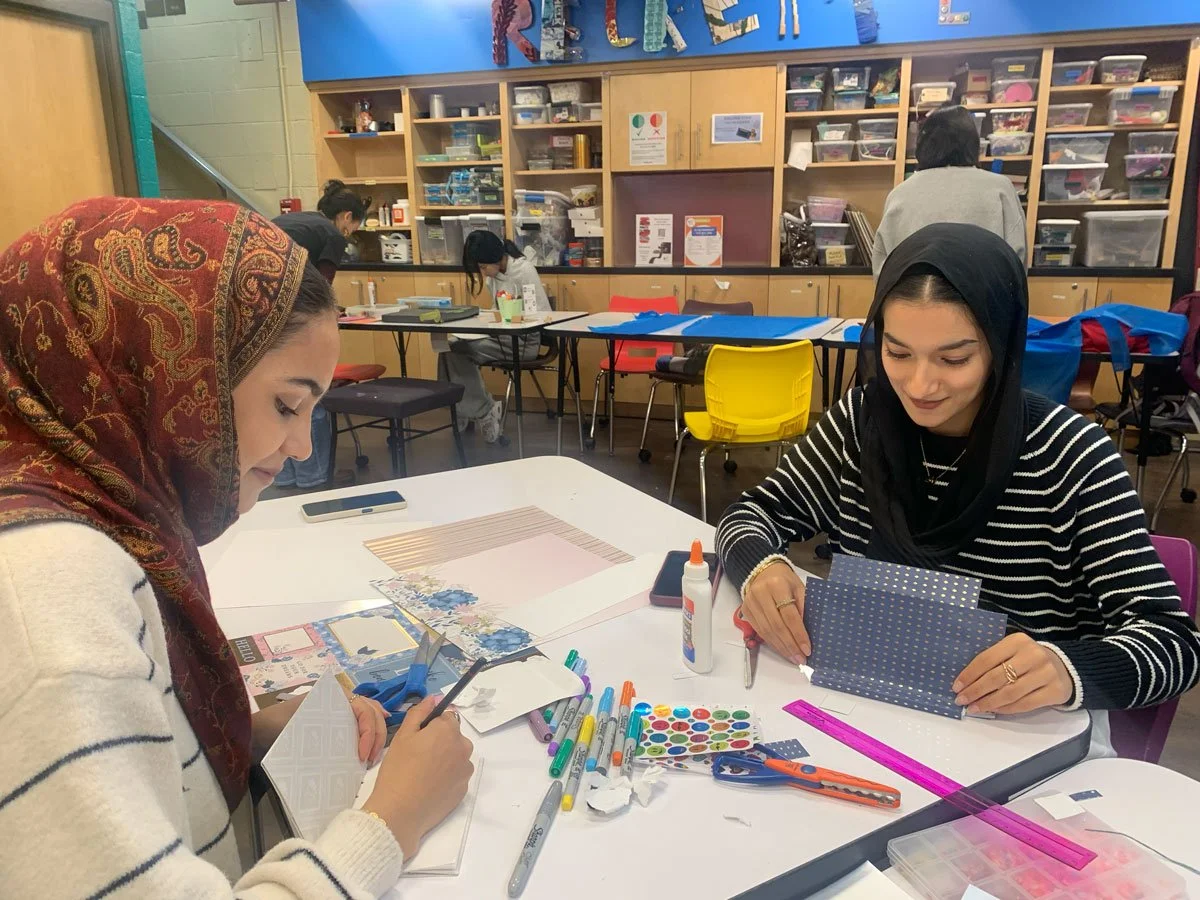 Two women sitting at table engaged in arts and crafts, surrounded by markers, glue, scissors, paper, and craft supplies, in a classroom or workshop setting.