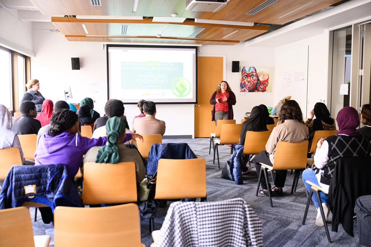 A woman is giving a presentation to a group of diverse people seated in a conference room. The room has a large screen showing a presentation slide and artwork on the walls.