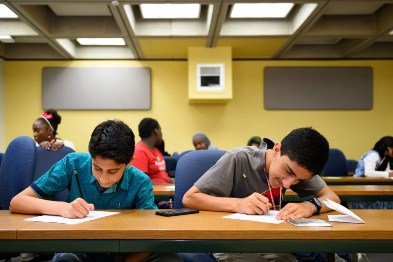 Two boys sitting at a desk and writing on paper during a classroom activity, other students in the background, yellow walls with speakers and ceiling panels.