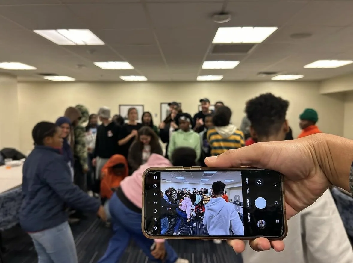Group of people gathered in a room, with someone taking a photo with a smartphone.