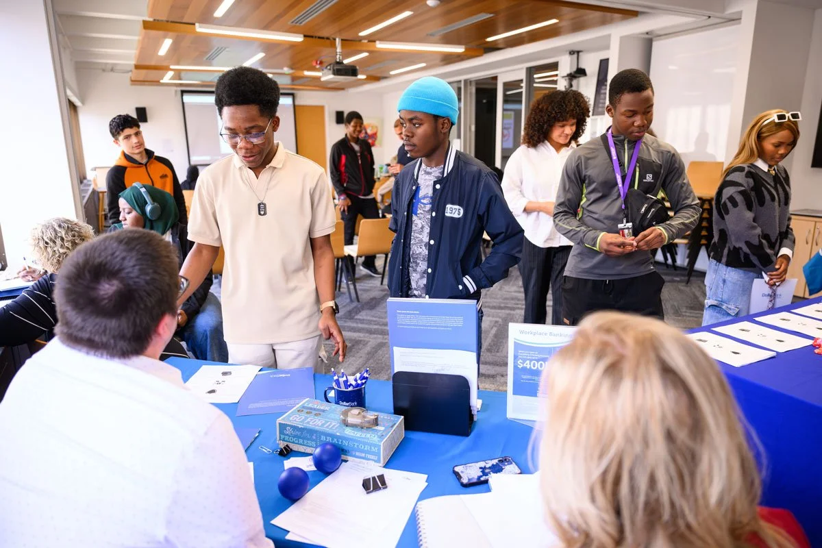 Group of diverse young people at an educational event or fair, engaging with representatives and exploring displays in a modern indoor setting.