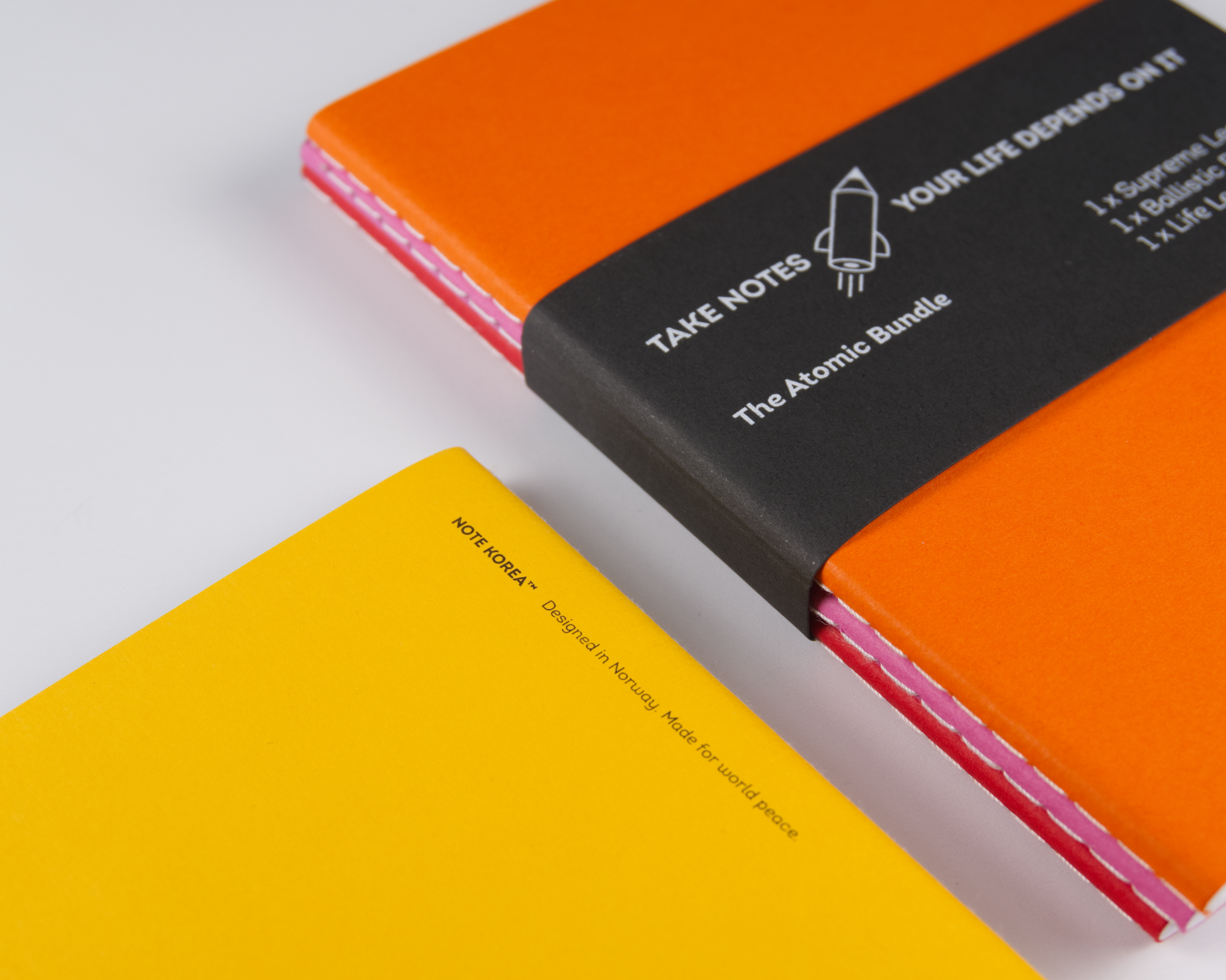 Close-up of colorful notebooks from Note Korea™, with one orange, one yellow, and a black one with white text and a rocket icon, on a white surface.