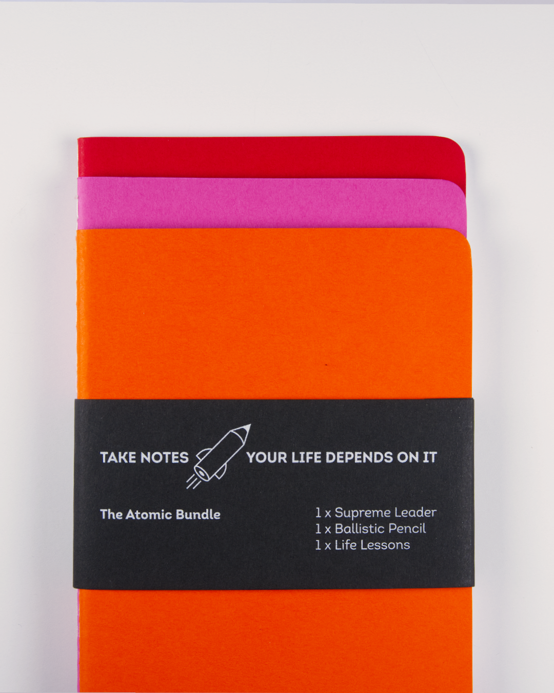 Colorful notebooks in red, pink, orange, and black with a black band reading, 'Take Notes, Your Life Depends on It,' accompanied by a rocket graphic and the text 'The Atomic Bundle' with items listed as Supreme Leader, Ballistic Pencil, Life Lessons.
