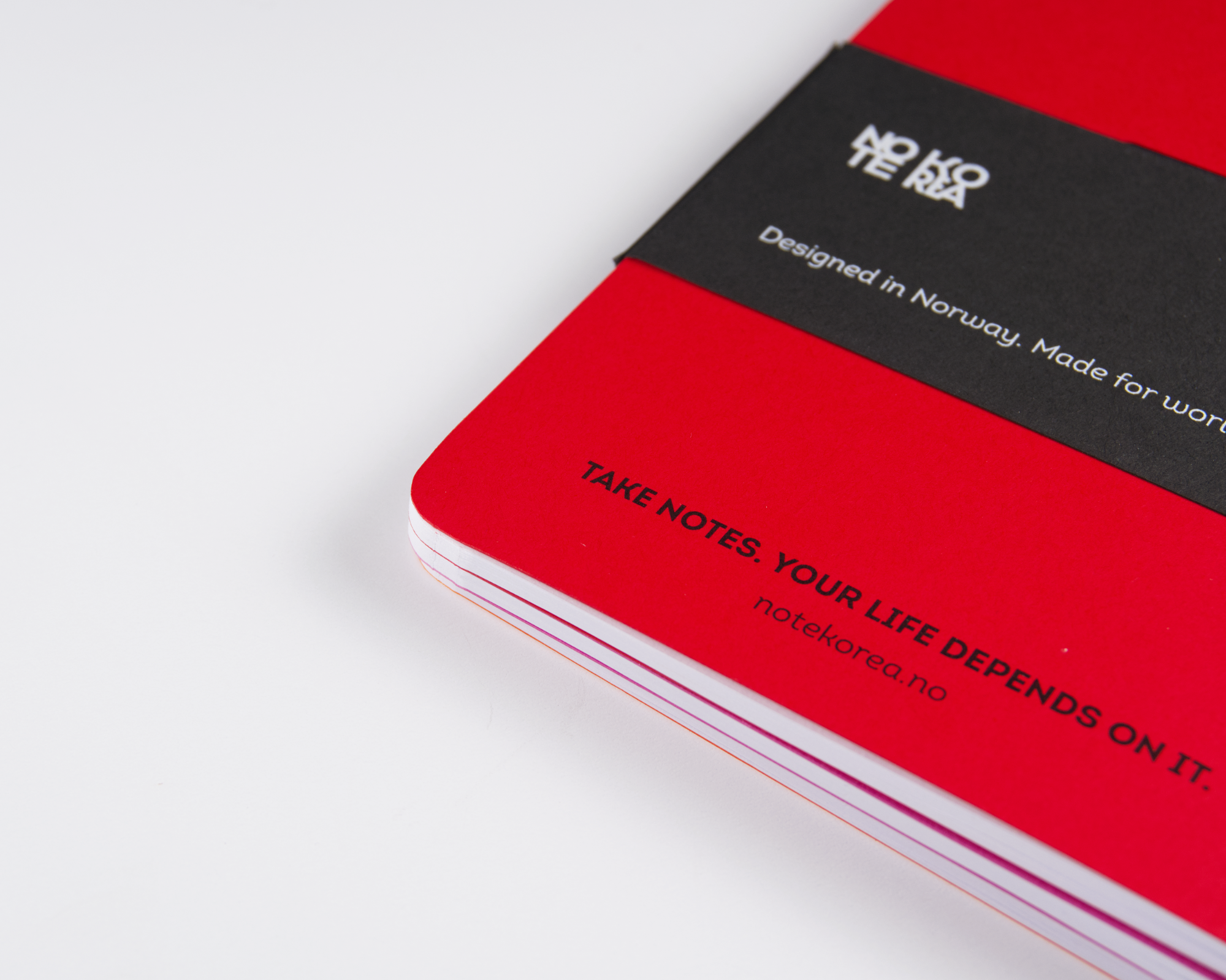 Close-up of a red Note Korea™ notebook with a black label that says 'Designed in Norway. Made for world peace' and partially visible text 'TAKE NOTES. YOUR LIFE DEPEND' on the cover.