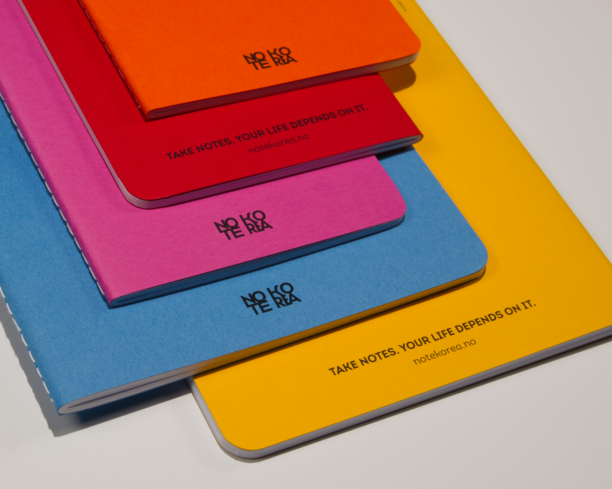 Three colorful notebooks in pink, orange, and blue stacked on top of each other, with the orange notebook on top, displaying the text "NO TE NO" and the pink notebook showing the website "notekorea.no." The yellow notebook underneath has the text "TA