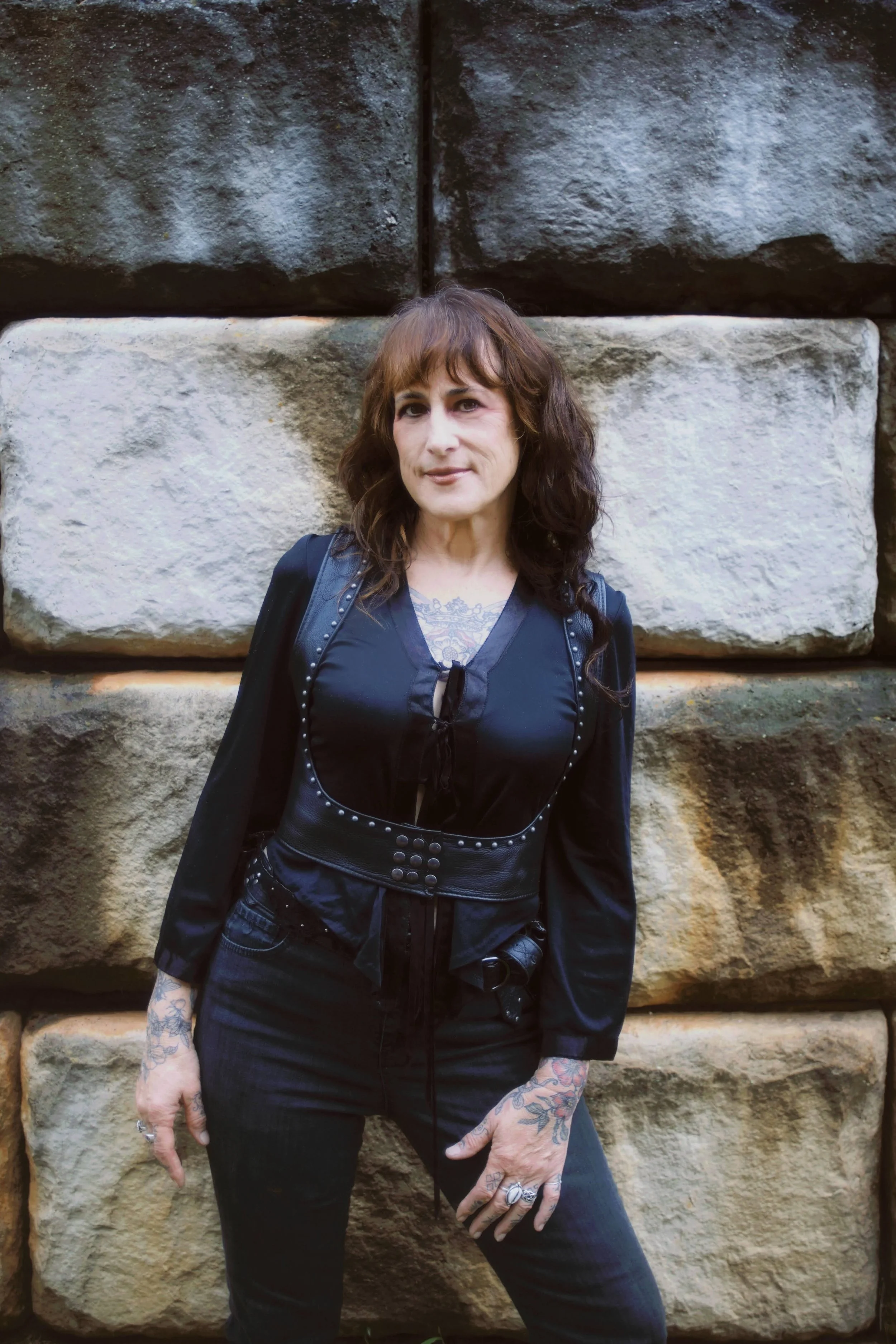 Woman with brown wavy hair and tattoos on her hands standing in front of large stone blocks.