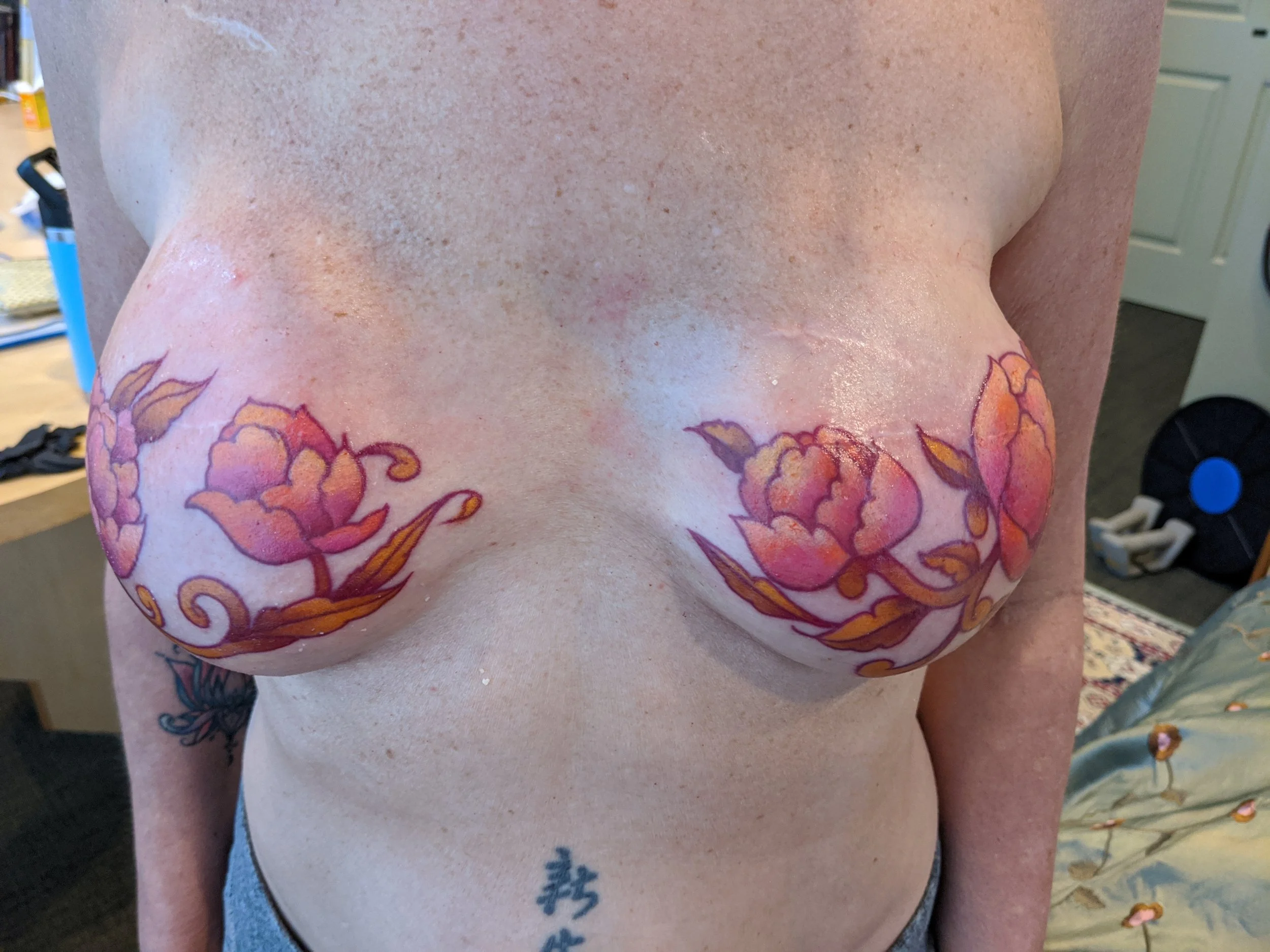 Close-up of a woman's bare chest with pink and orange floral tattoos on her breasts.