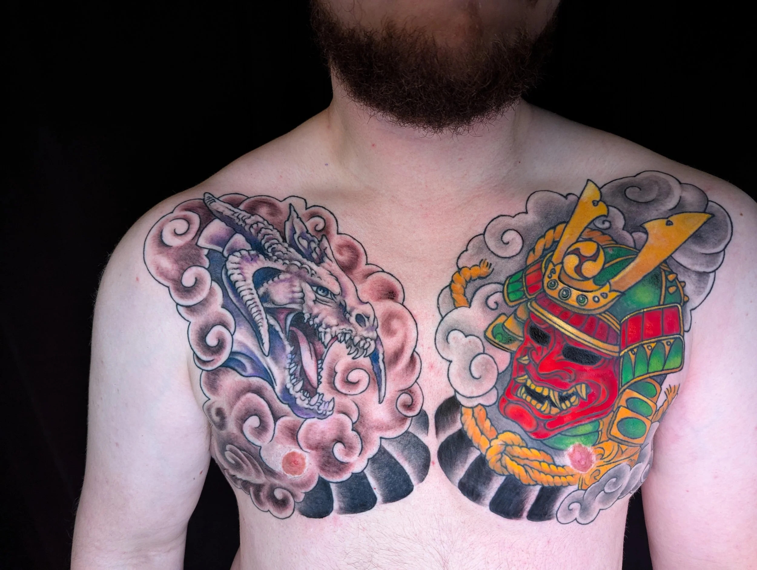Colorful tattoo on a person's chest featuring a fierce dragon on the left side and a traditional Japanese mask on the right side, both surrounded by swirling clouds.