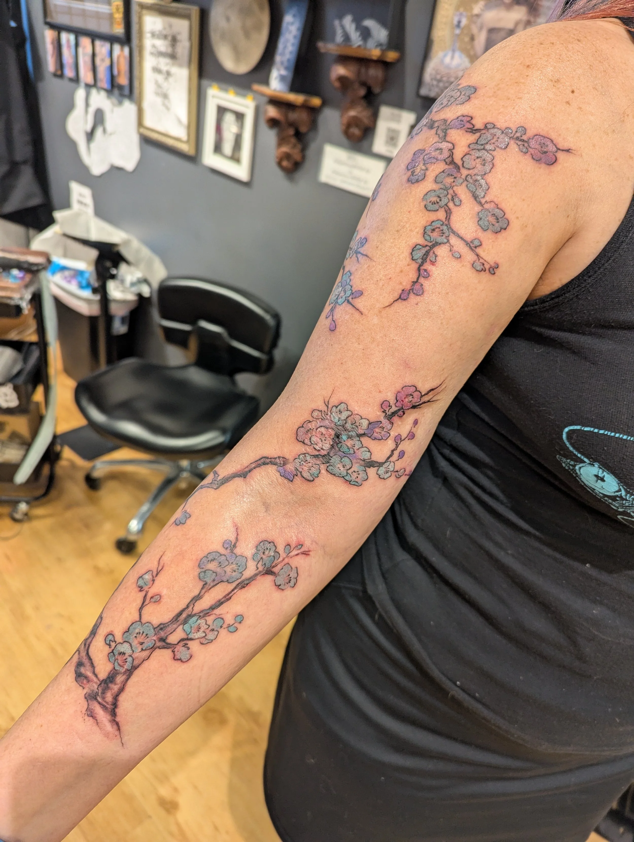A person with a new tattoo of cherry blossoms on their upper arm and forearm. The tattoo features pink and purple flowers with detailed black outline.