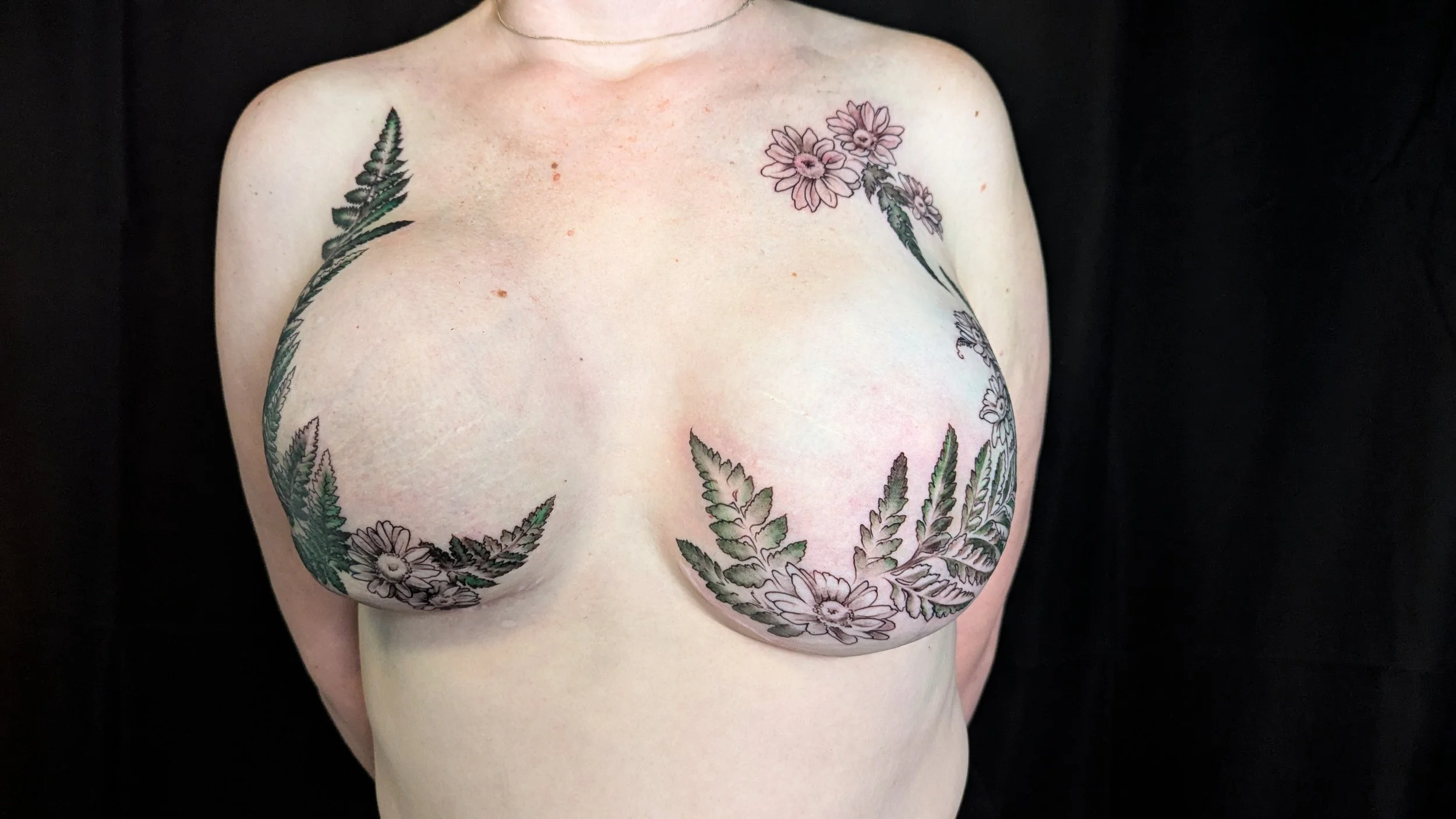 A person with a floral tattoo on their chest and shoulders. The tattoo features black and green leaves and pink flowers, arranged in a circular pattern around the chest.