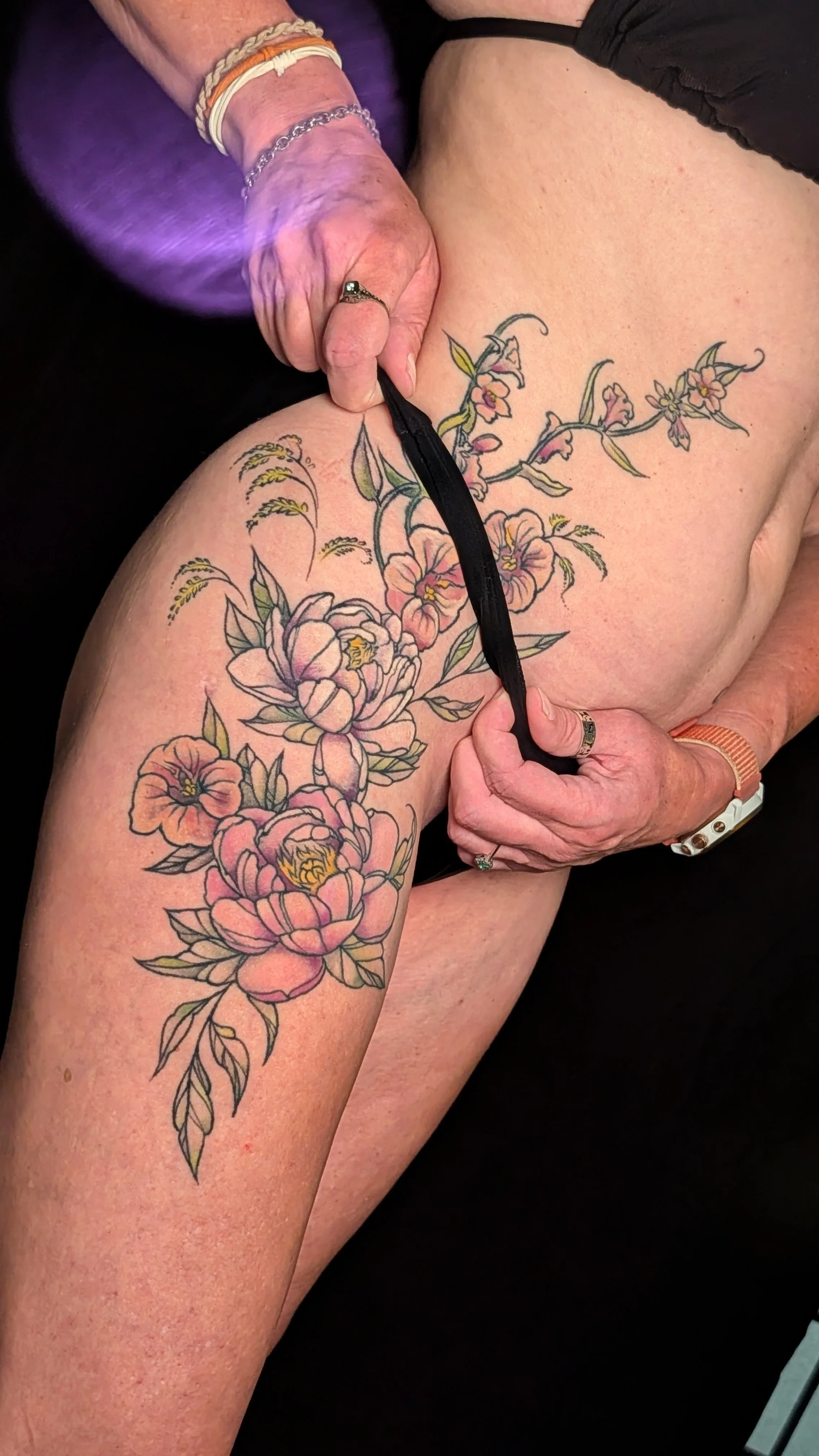 A person is holding a black ribbon or cloth, wearing multiple bracelets and a ring, near a large floral tattoo on their thigh. The tattoo features pink and peach flowers with green leaves and vines.