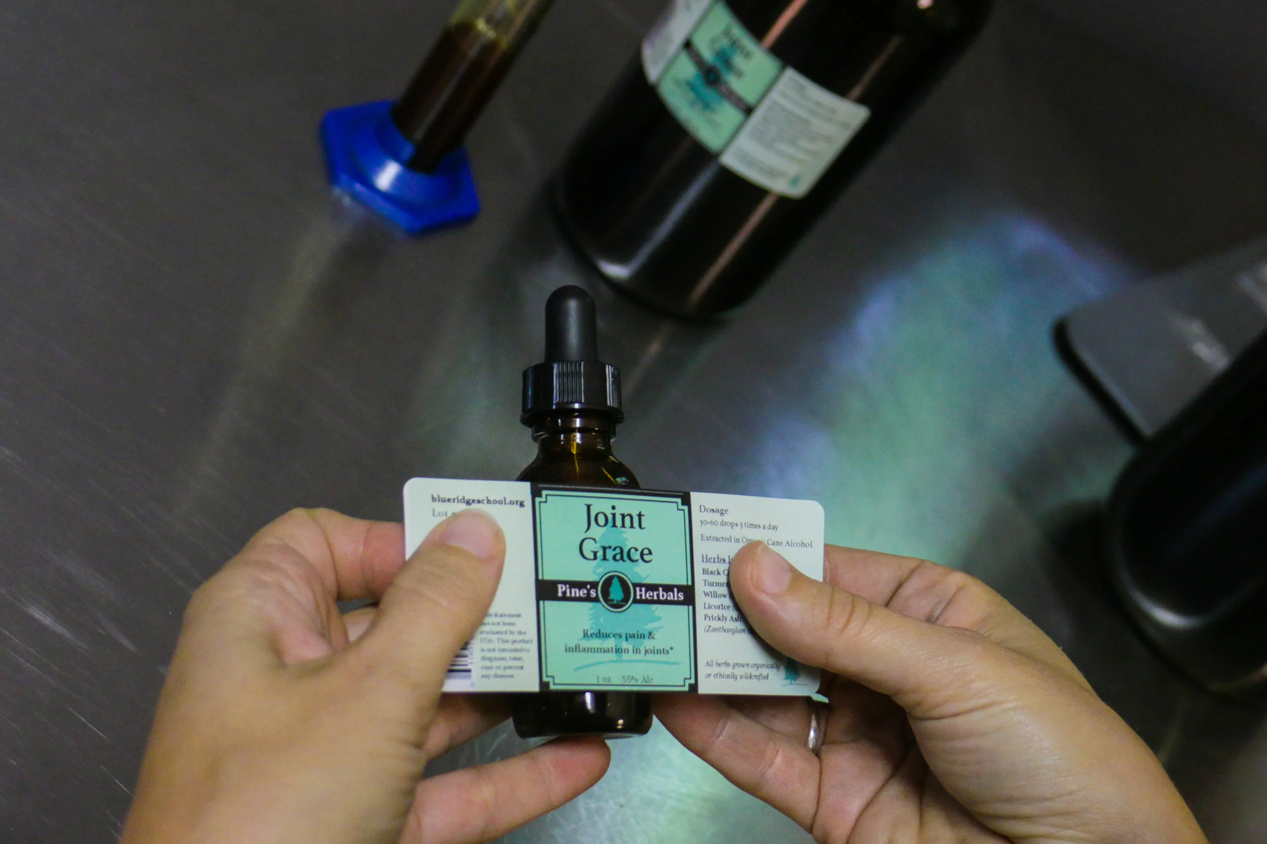 Two hands applying the Pine's Herbal Joint Grace tincture label to a 20x bottle in a lab