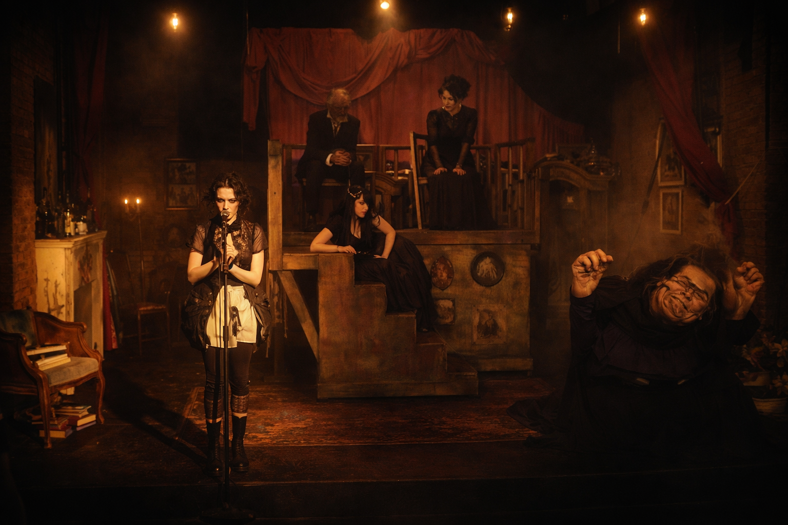 A theatrical scene with a woman singing into a microphone, four other actors on stage in dark costumes, and a set resembling an old house interior with red curtains and vintage furniture, including a piano and a bookshelf.