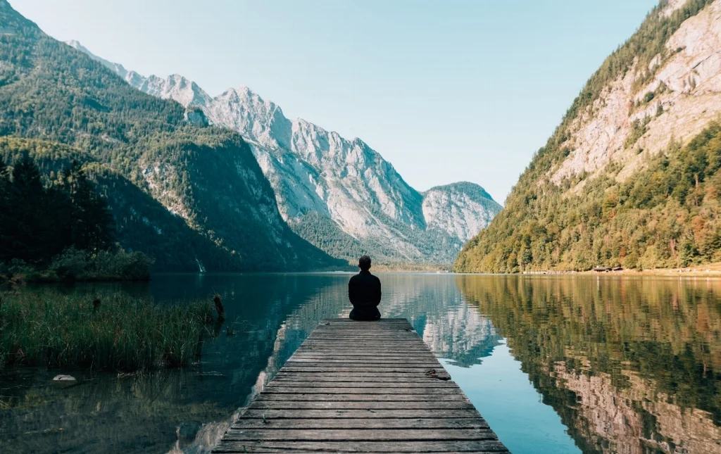 Man sitting on a wooden dock on a lake with mountains reflected in the lake’s surface.