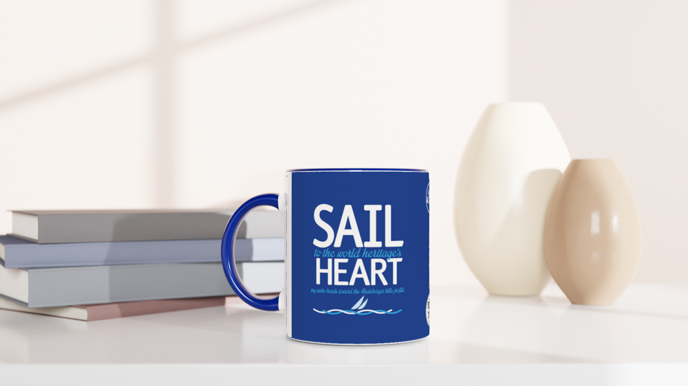The picture shows a left side view of the 11oz ceramic mug "SAIL TO THE WORLD HERITAGE HEART" from the series HIGH COAST SAILOR #TAGMUG™ with blue color inside on a desktop