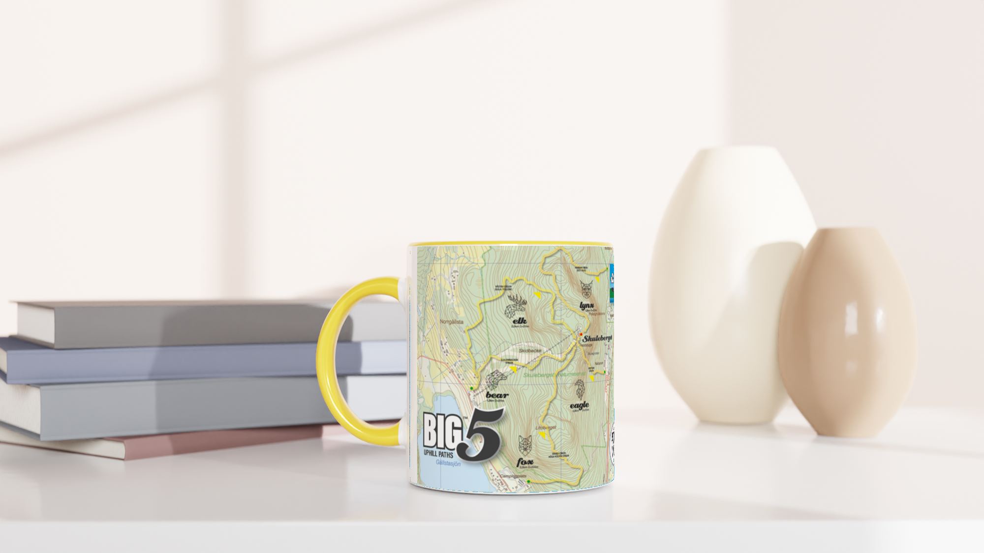 The picture shows a left side view of the 11oz ceramic mug "SKULEBERGET ELK PATH" from the series “SKULE BIG5" #TAGMUG™ with yellow color inside on a desktop