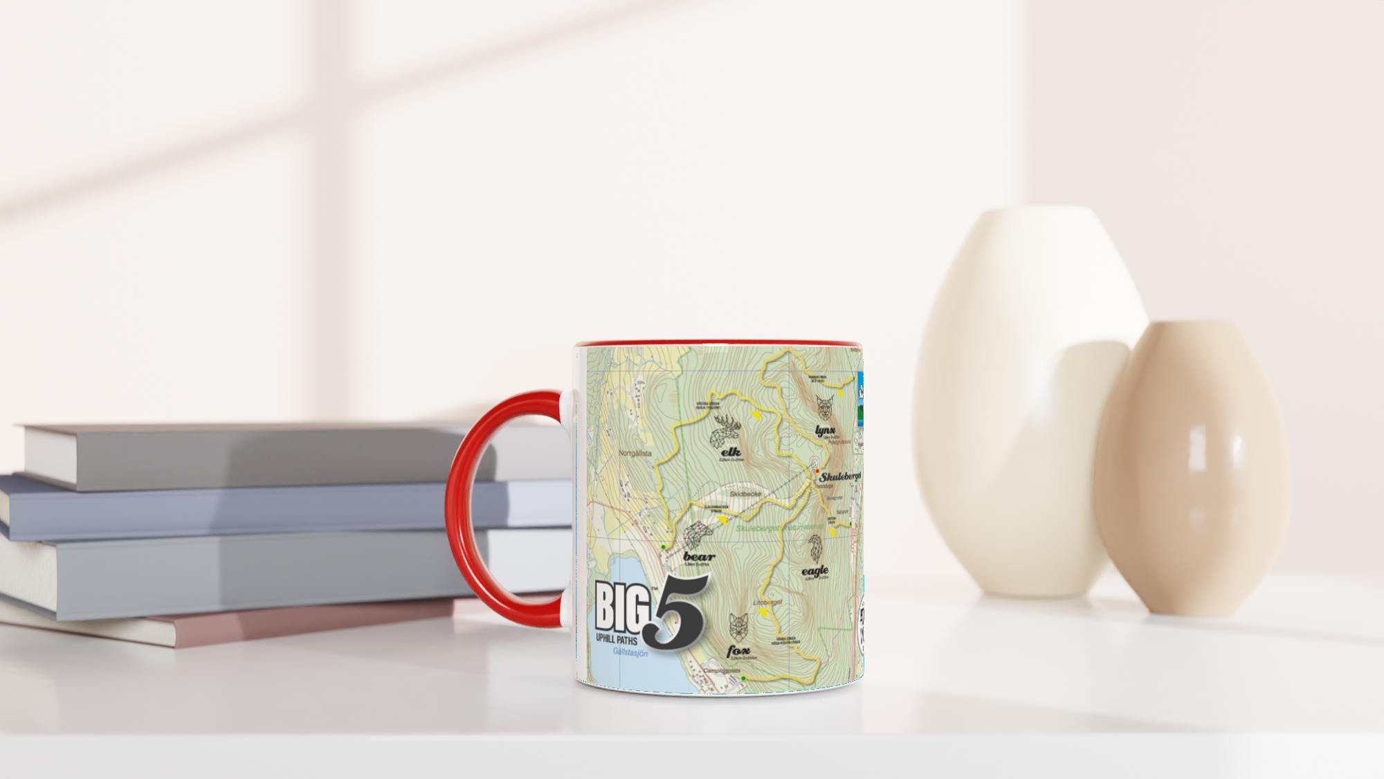 The picture shows a left side view of the 11oz ceramic mug "SKULEBERGET BEAR PATH" from the series “SKULE BIG5" #TAGMUG™ with red color inside on a desktop
