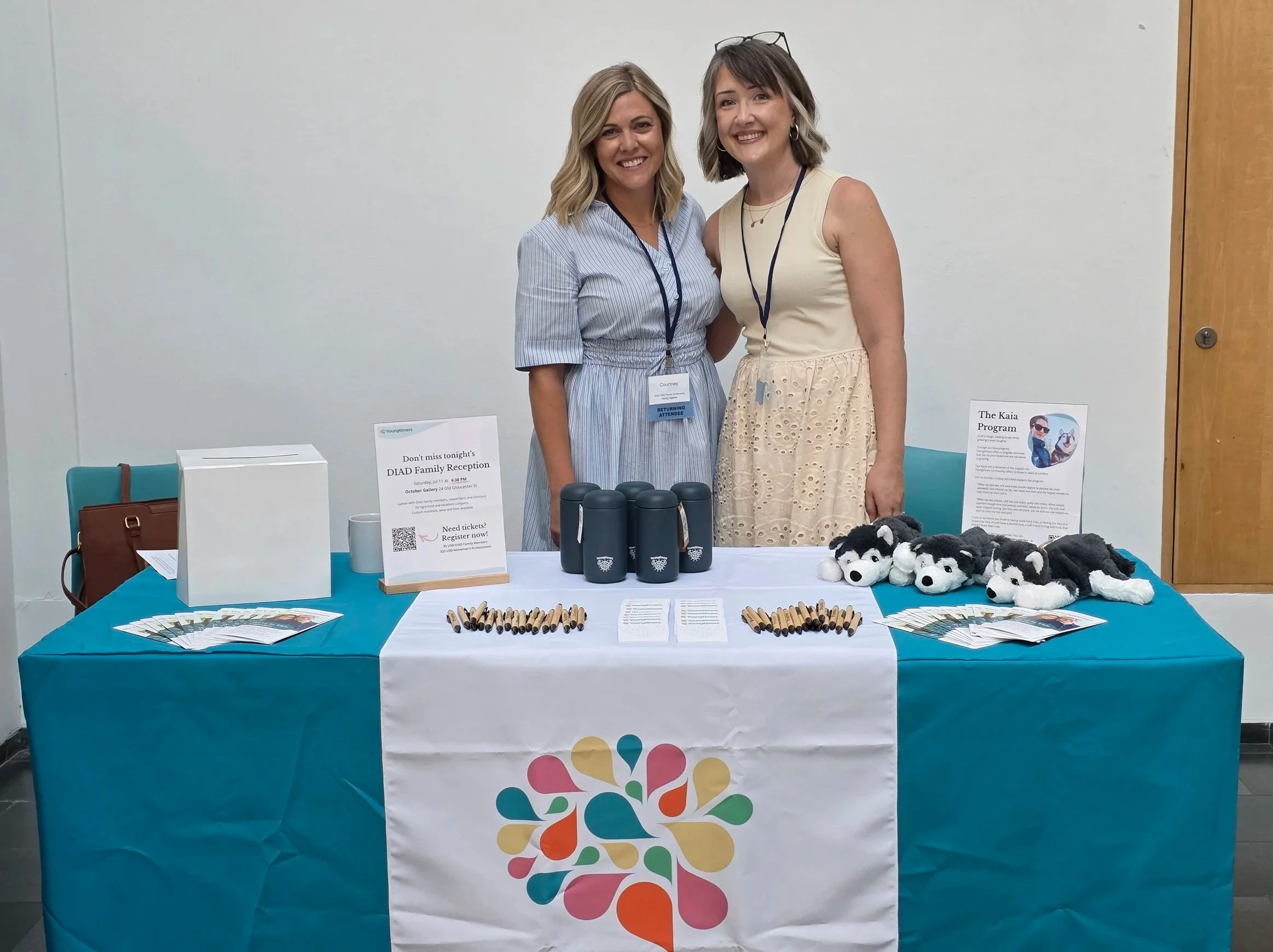 Two women smiling behind a Youngtimers event table with informational materials, a Kaia Program sign, and plush husky toys, promoting a DIAD Family Reception