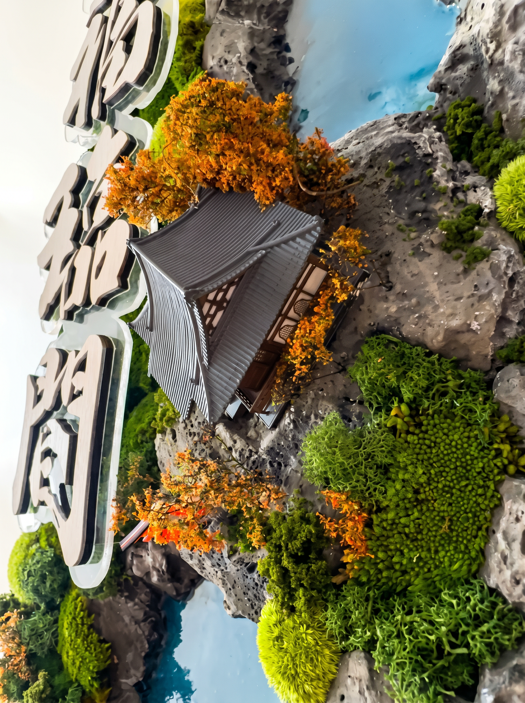  A Japanese-inspired preserved moss installation featuring a miniature temple nestled within lush landscapes, accented by handcrafted resin water bodies that mimic natural ponds and vibrant autumn trees that beautifully complement the warm wood inter