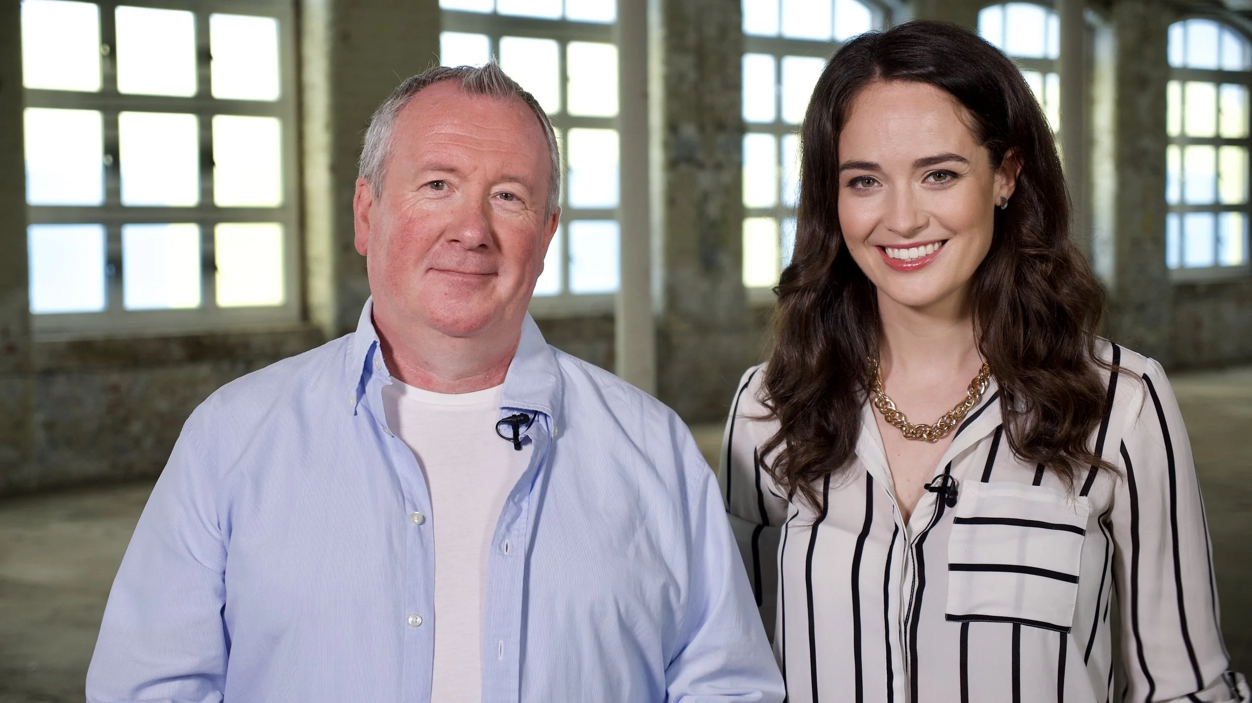 A middle-aged man with gray hair and a woman with long curly dark hair standing side by side in a spacious room with large windows and brick walls,
Promo photo for The People's History Show at Up Next Studios Glasgow