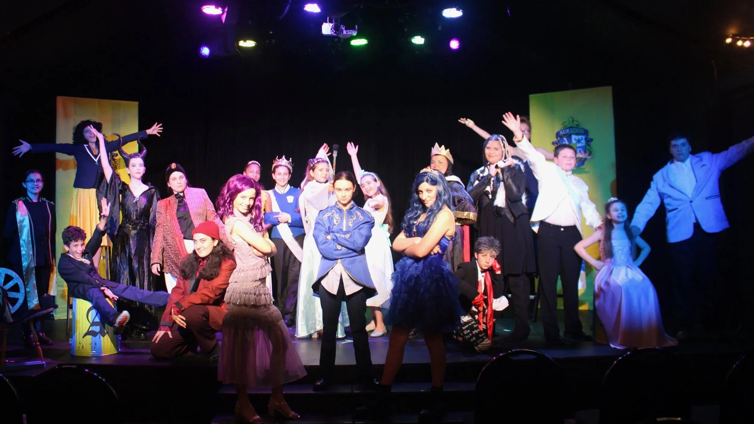 Group of children and teenagers on stage during a theatrical performance, dressed in costumes representing various characters, with some wearing crowns and others in fantasy or historical attire, under colorful stage lighting.