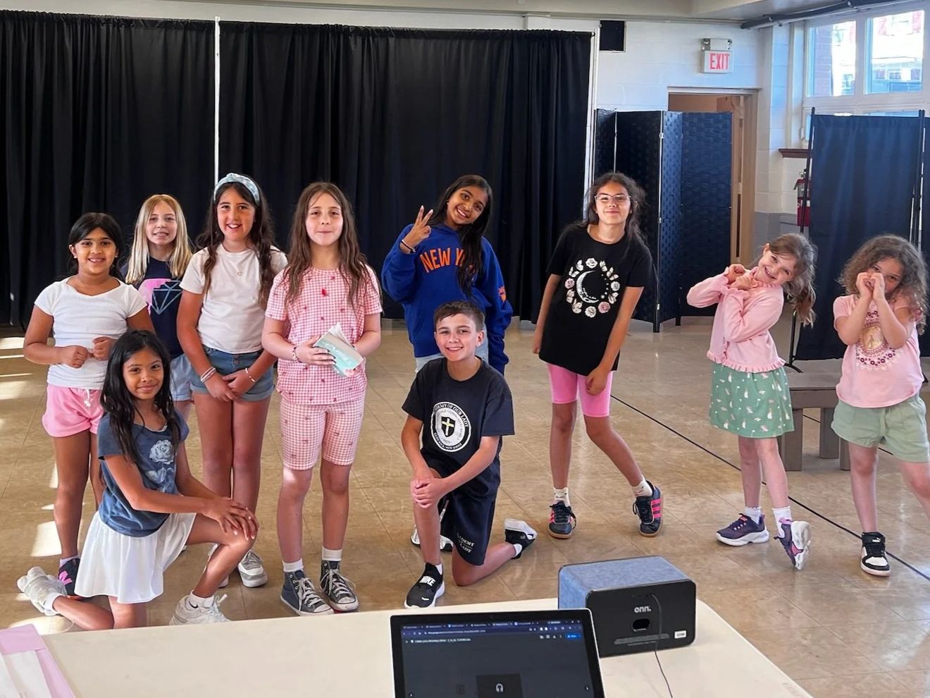 Group of ten diverse children posing and smiling in a school auditorium, some standing and some kneeling. Behind a black curtain and near a table with a tablet and speaker, with windows and an exit sign in the background.