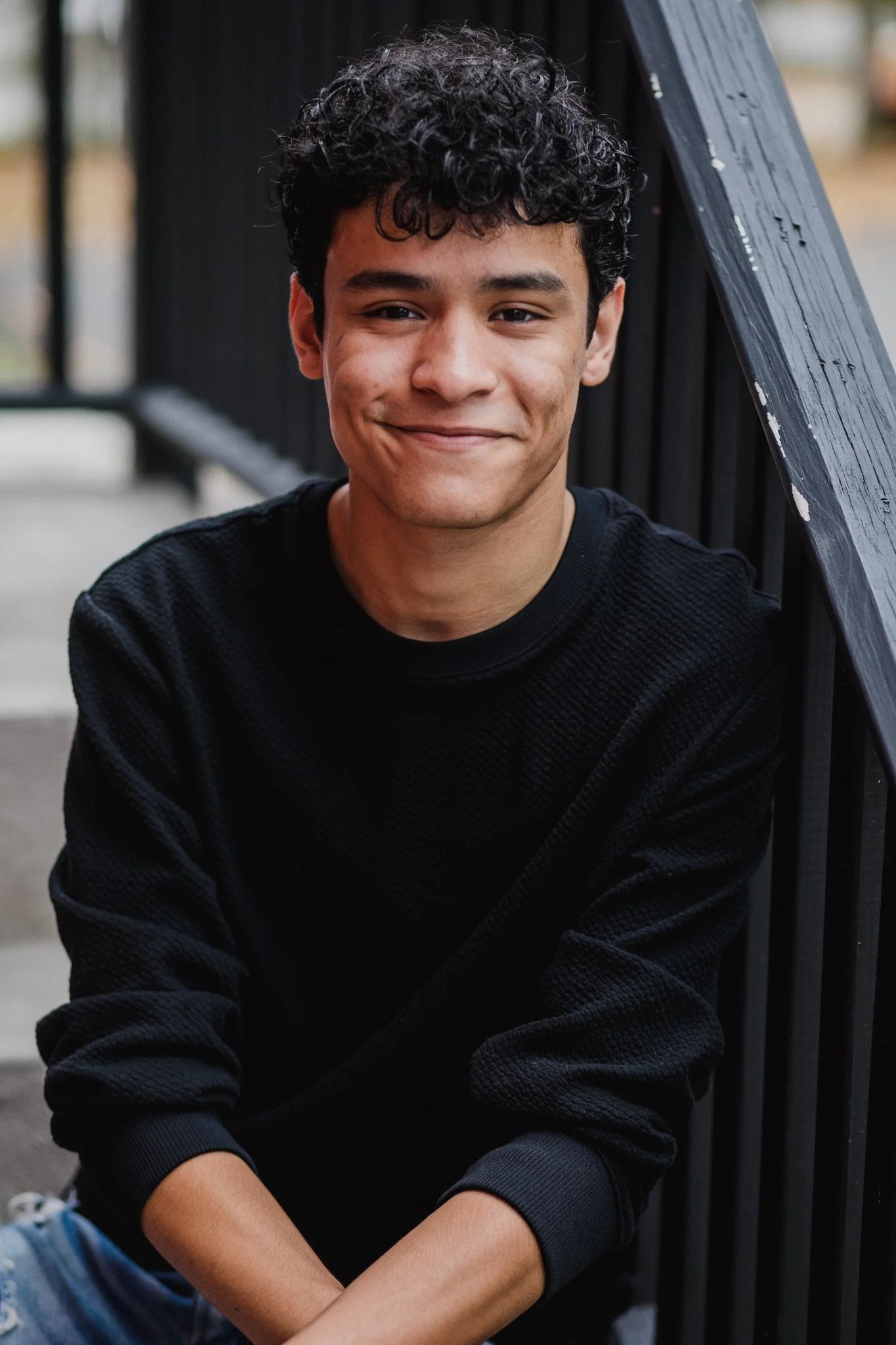 A young man with curly hair smiling and sitting near a black fence on an outdoor staircase.