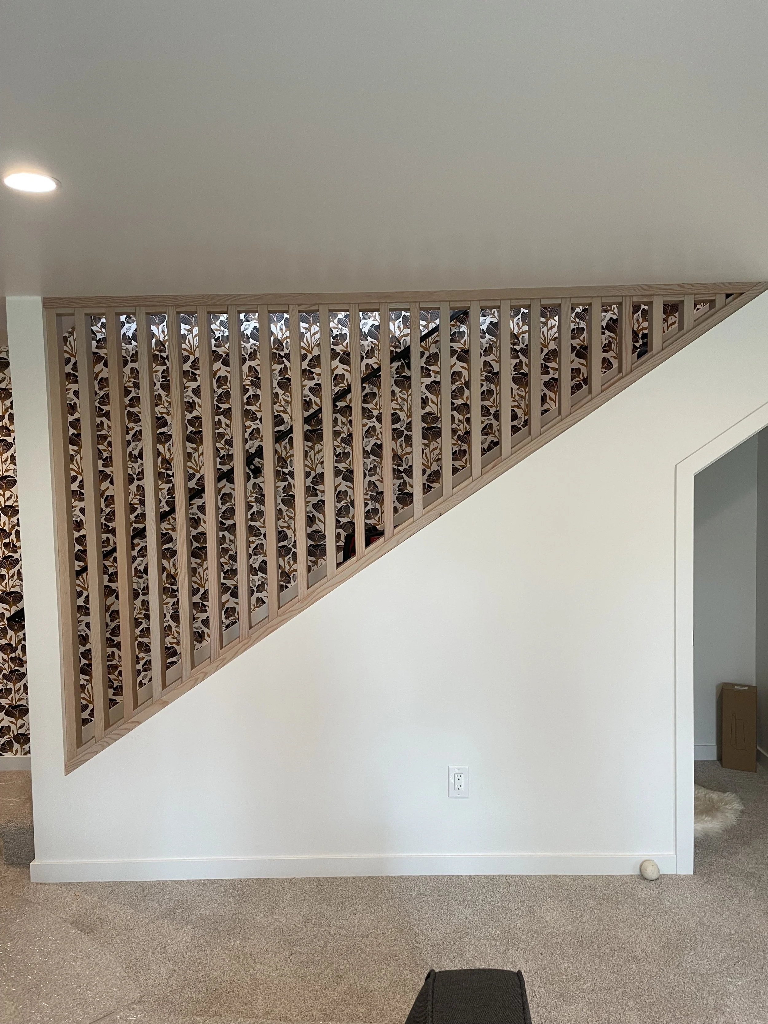 Interior view of a staircase with a wooden railing, white and patterned wallpaper, light beige carpet, and an electrical outlet on the wall.