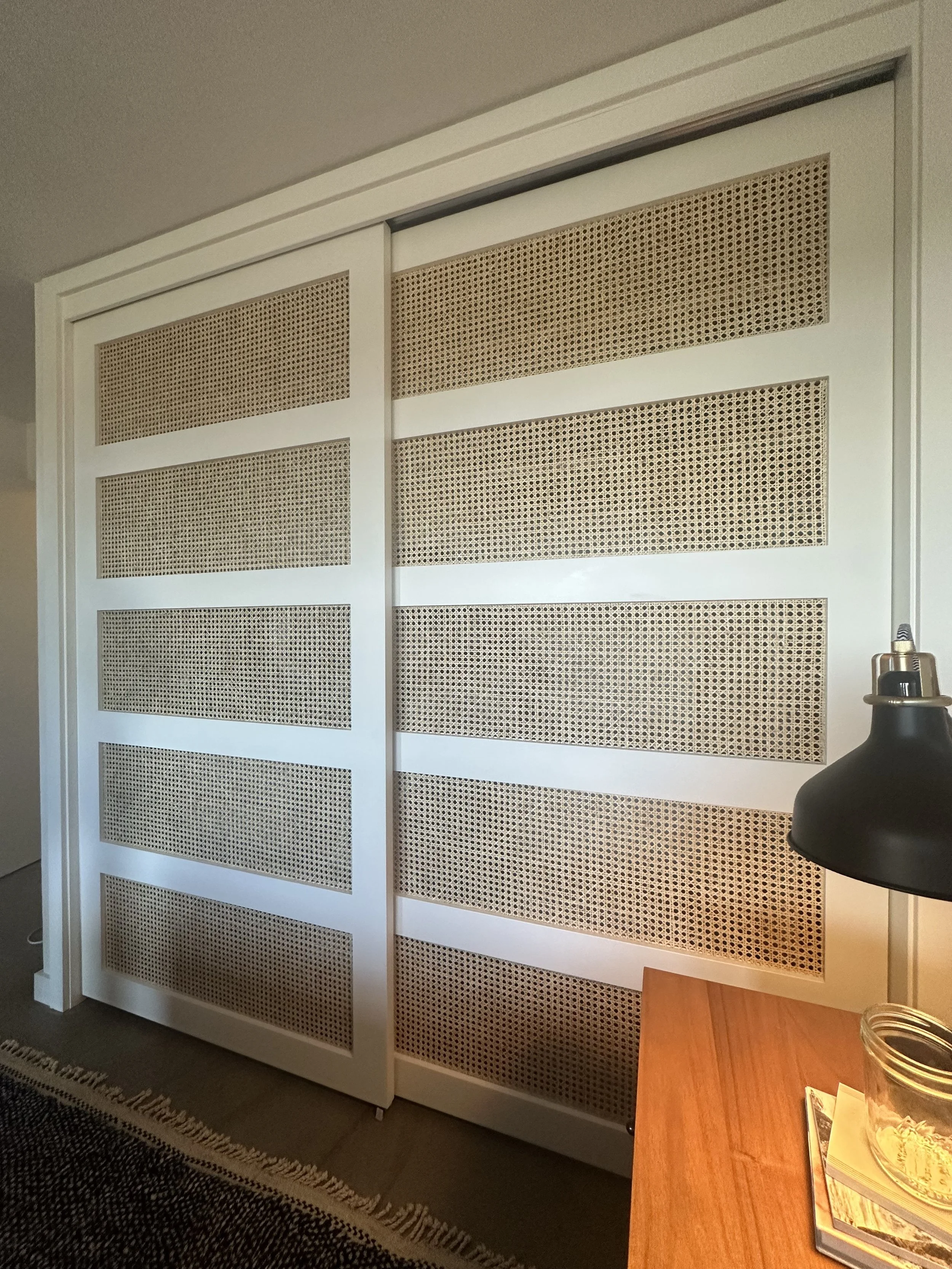 Sliding closet doors with a white frame and woven cane panels, partially open, in a room with a wooden desk, black lamp, and a glass jar.