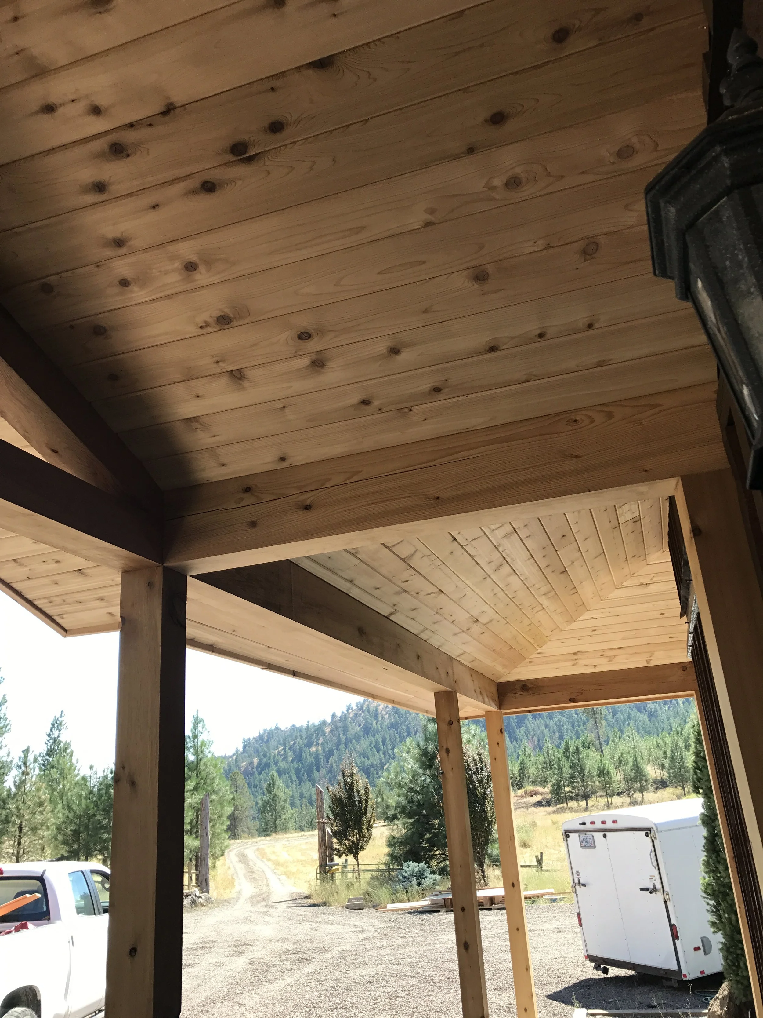 View of the underside of a wooden porch roof with wooden beams and natural scenery in the background, including trees, a dirt road, and a trailer.