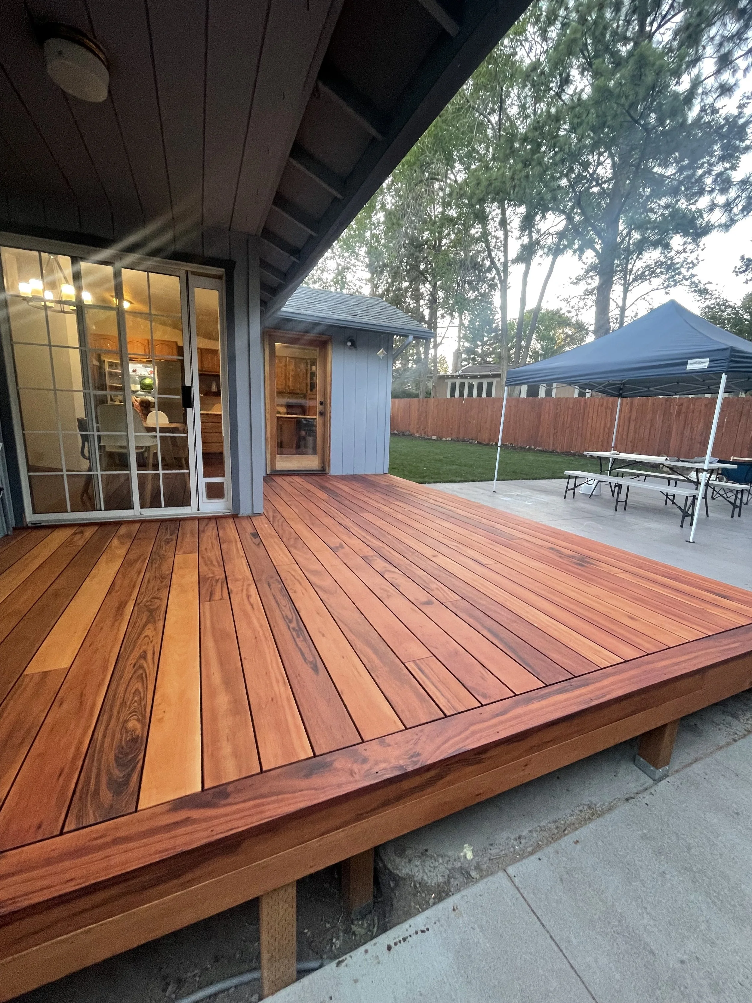 A backyard with a wooden deck attached to a house, glass sliding door, there are outdoor tables and a canopy in the yard, and a fenced backyard with trees.