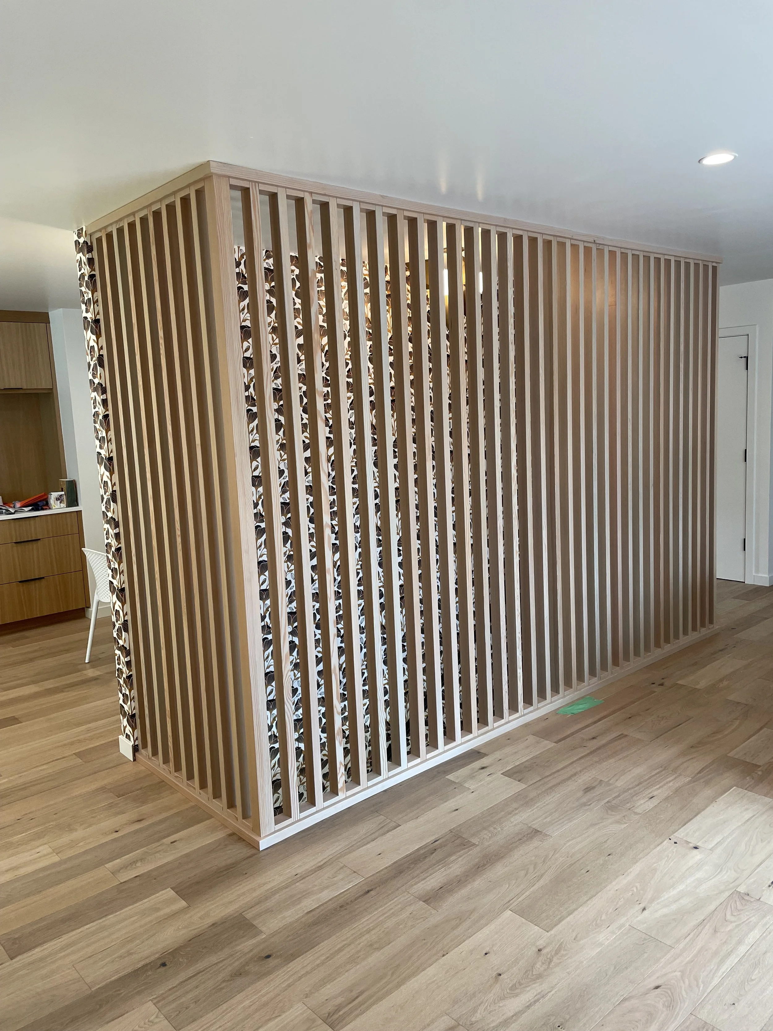 A room divider made of vertical wooden slats with a floral patterned fabric or wallpaper behind it, separating part of a living space with wooden flooring and some furniture.