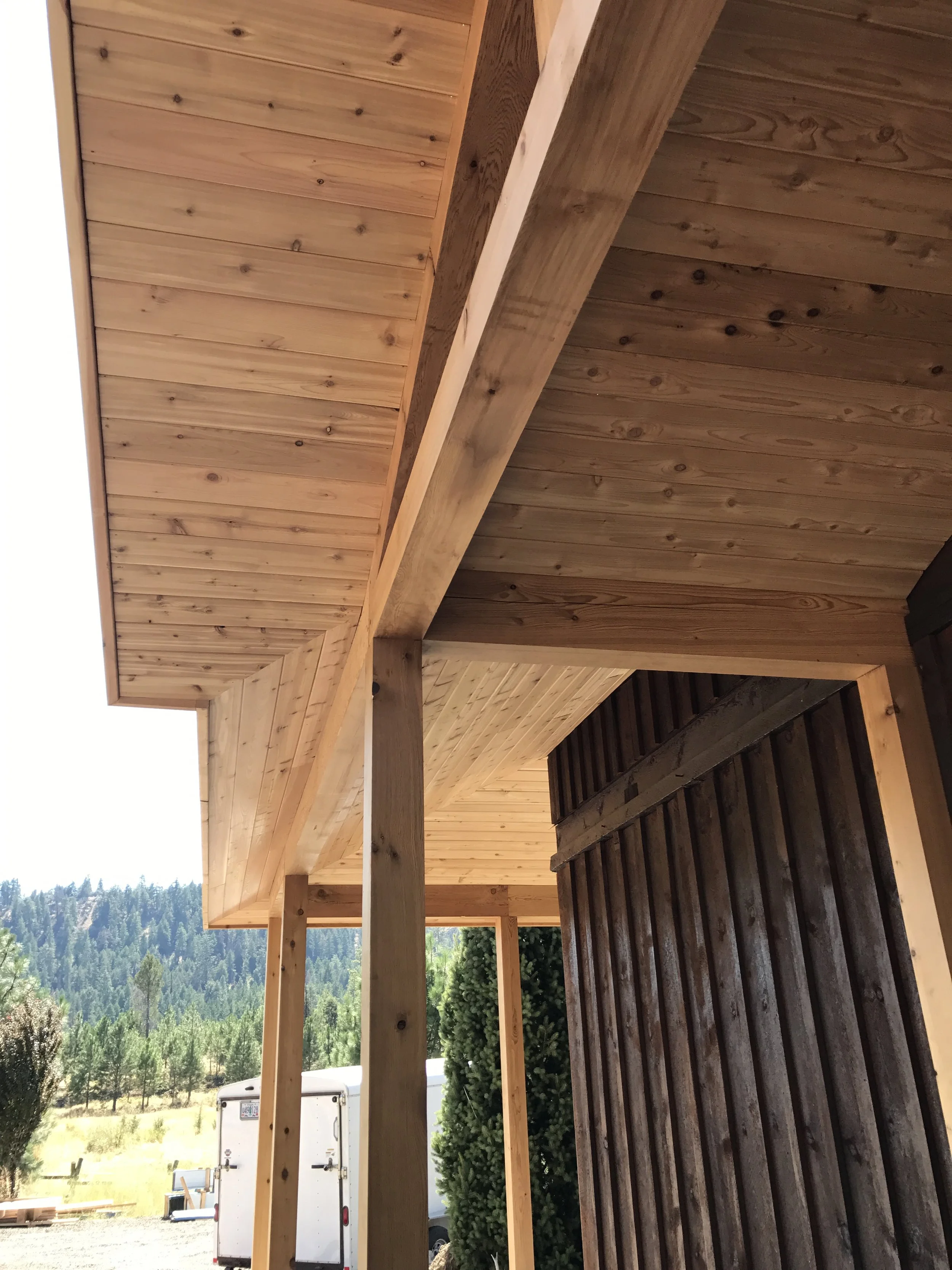 View of a wooden porch with a wooden roof, supported by wooden beams, with a forested mountain landscape in the background.