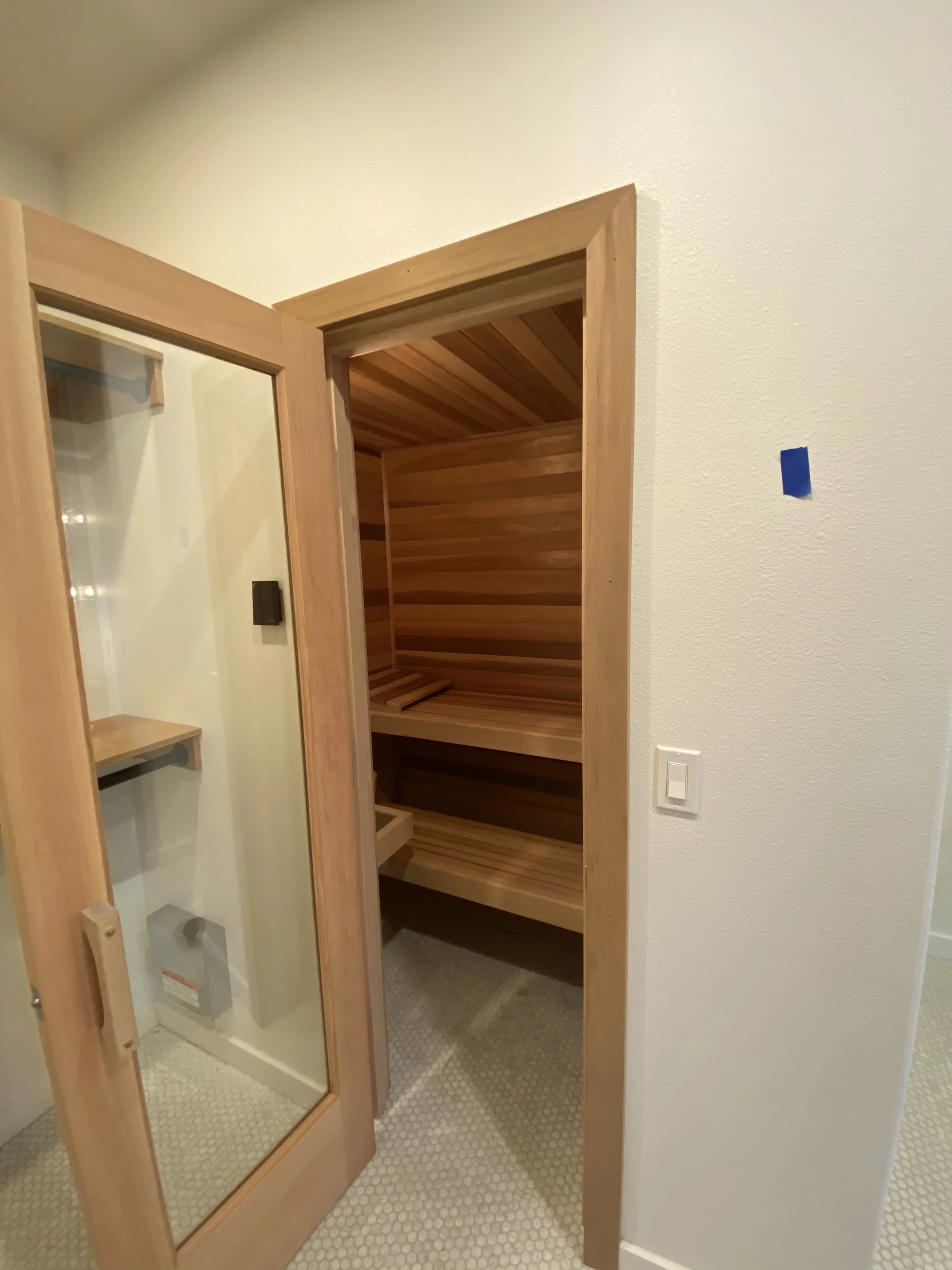 Interior of a small wooden sauna with shelves and seating, showing a door with a glass panel.