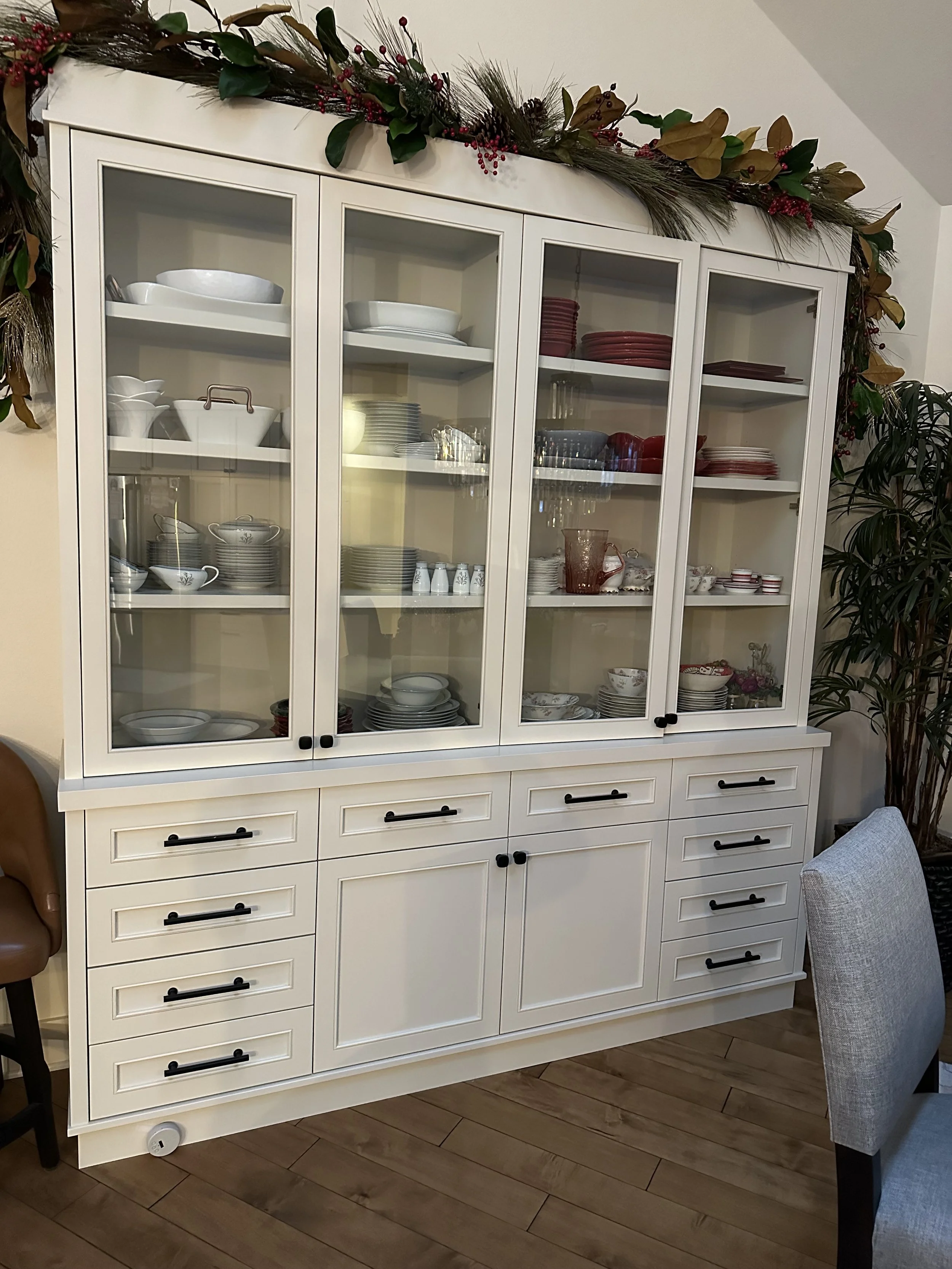 A white china cabinet with glass doors displaying various dishes and glassware, topped with holiday greenery and red ornaments in a dining room.